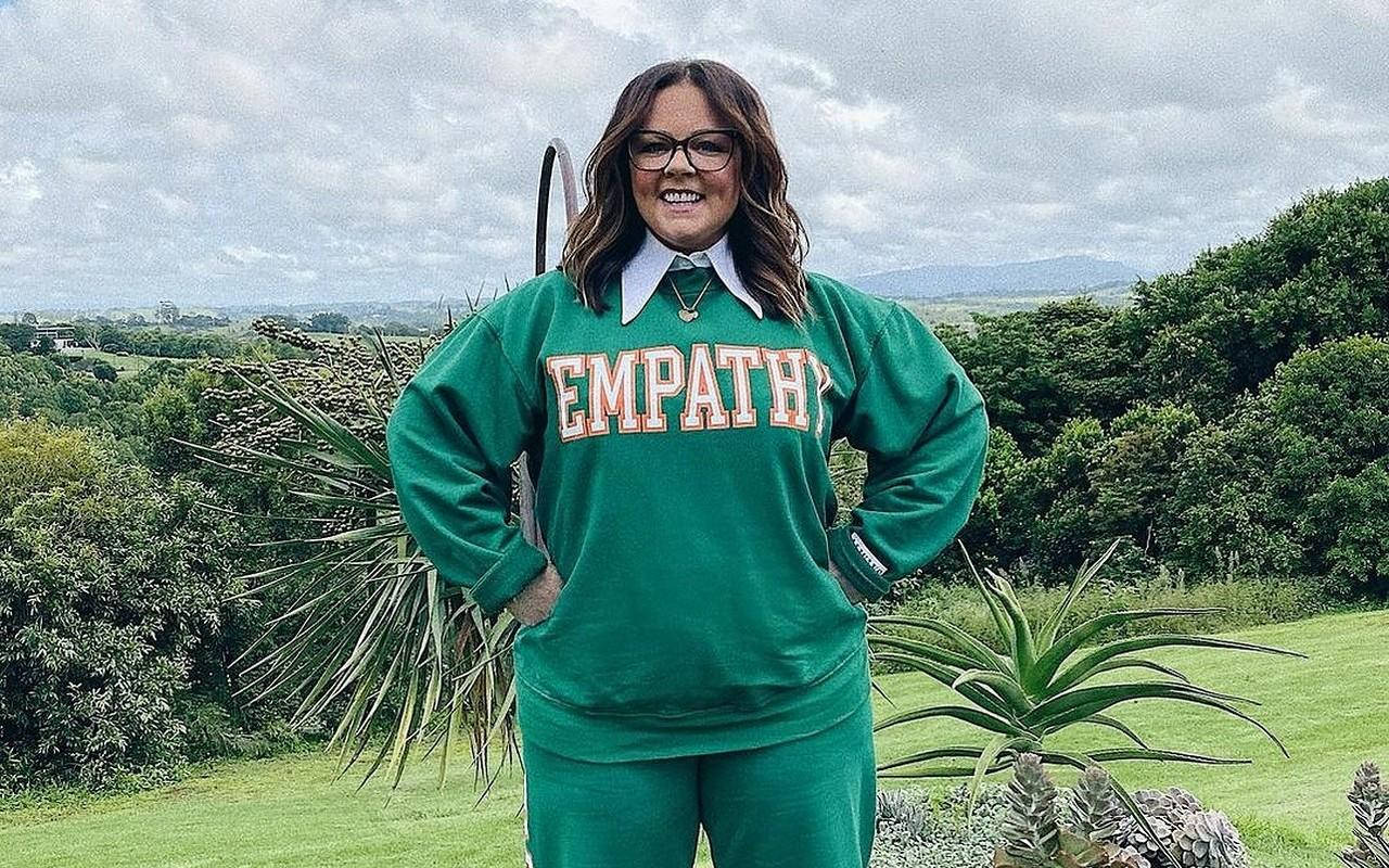 Celebrity 桌布 壁紙 — 一位微笑的女性穿著綠色 'EMPATHY' 運動衫，自信地站在長滿青草的山丘上，周圍有茂密的樹木和遠處的山脈，天空多雲。