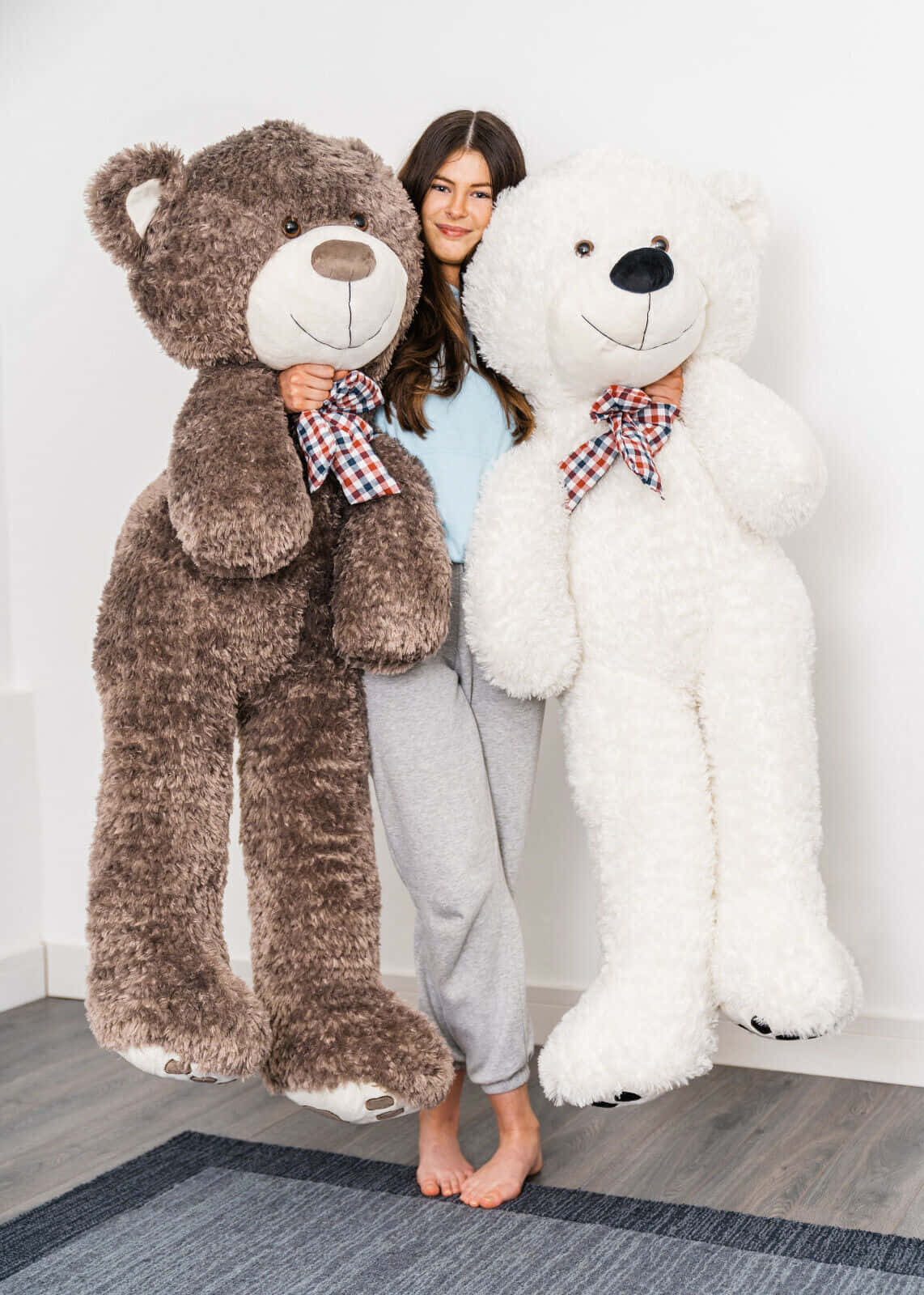 Teddy Bear 桌布 壁紙 — 一位微笑的女性穿著舒適的家居服，站在兩隻巨大的絨毛熊之間，一隻棕色和一隻白色，兩隻都戴著格子蝴蝶結。