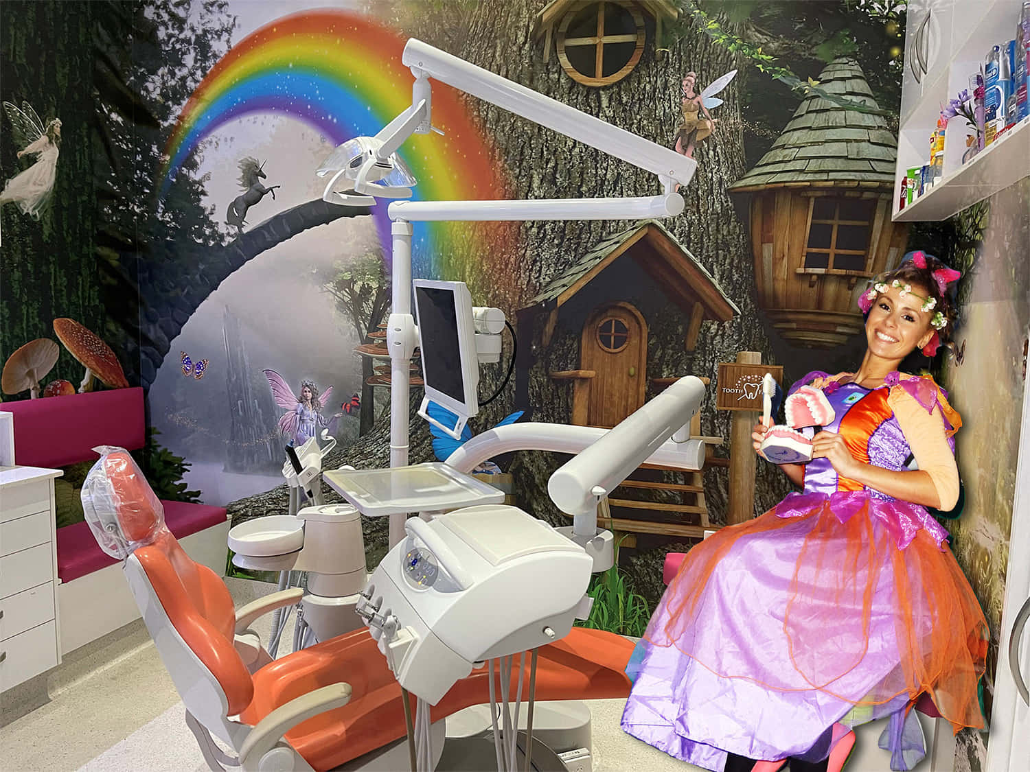 Tooth Fairy 桌布 壁紙 — 一位微笑的女性穿著色彩繽紛的公主服裝，坐在現代牙科椅旁，背景是奇幻的森林壁畫，有彩虹和仙女屋。