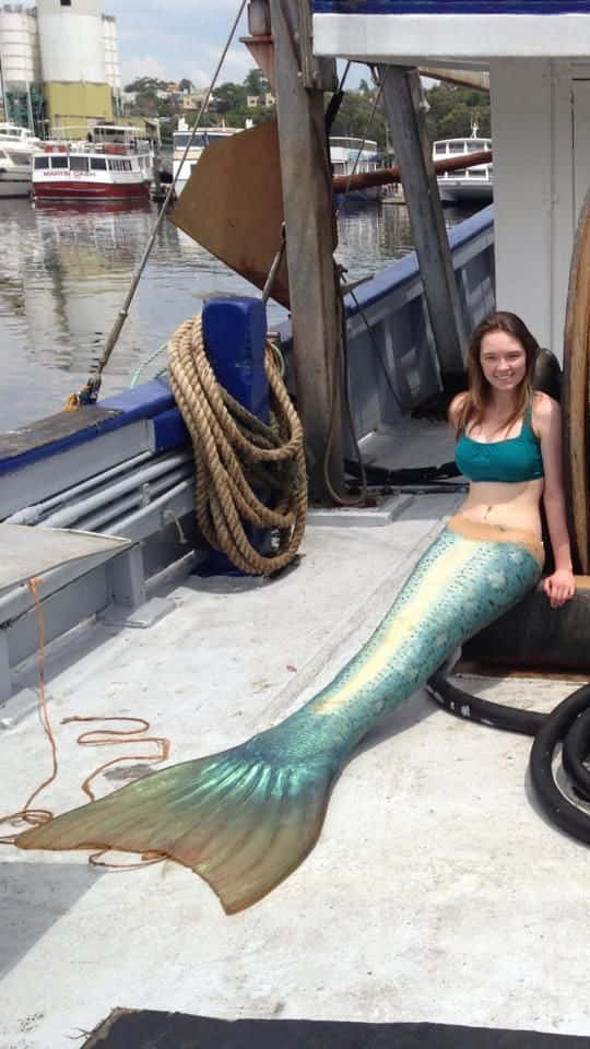 Mermaid Real Life 桌布 壁紙 — 一位微笑的女性穿著藍綠色美人魚尾巴，在一個停泊在港口的風化船上擺姿勢，背景有其他船隻和建築物。