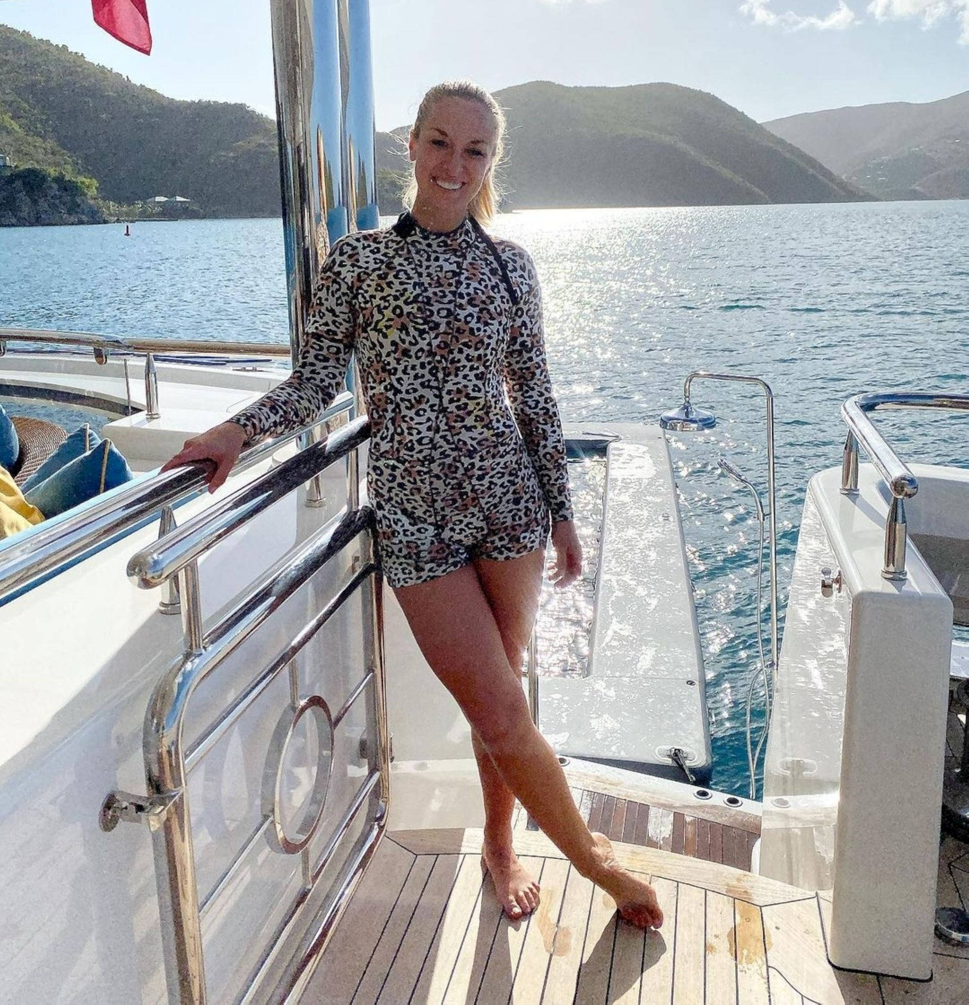 Yacht 桌布 壁紙 — 一位微笑的女性穿著豹紋泳衣，靠在遊艇欄杆上，身後是碧綠的海水和翠綠的山丘，在明亮的陽光下。