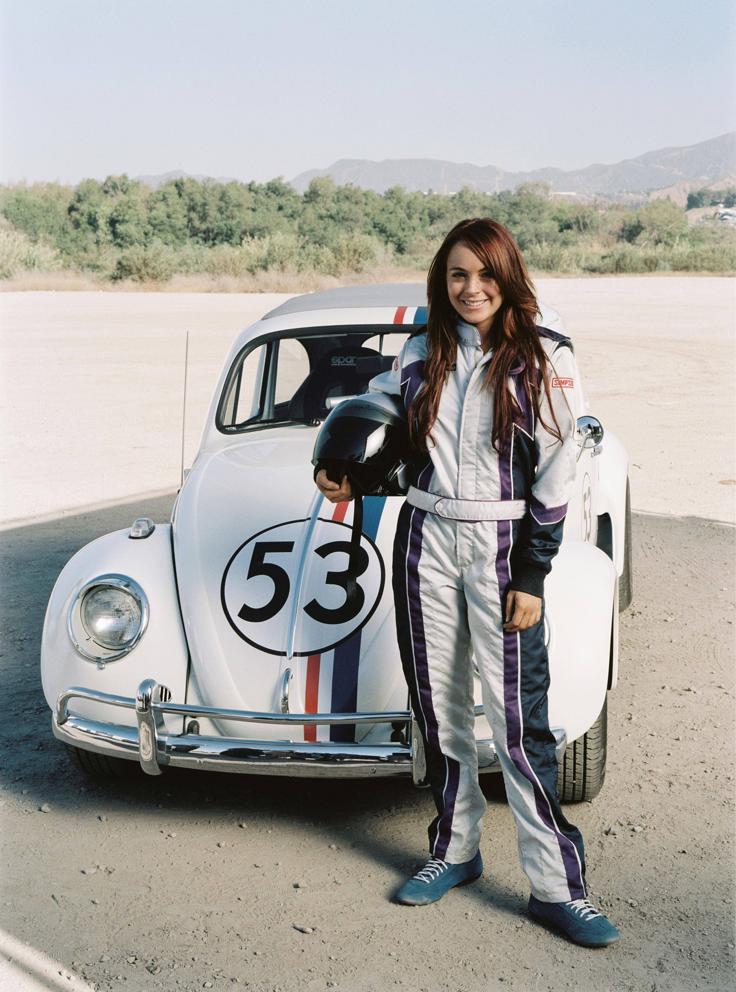 Herbie 桌布 壁紙 — 一位微笑的女性穿著賽車服，驕傲地站在一架白色復古賽車旁，車上標有53號，背景是陽光明媚的飛機跑道。