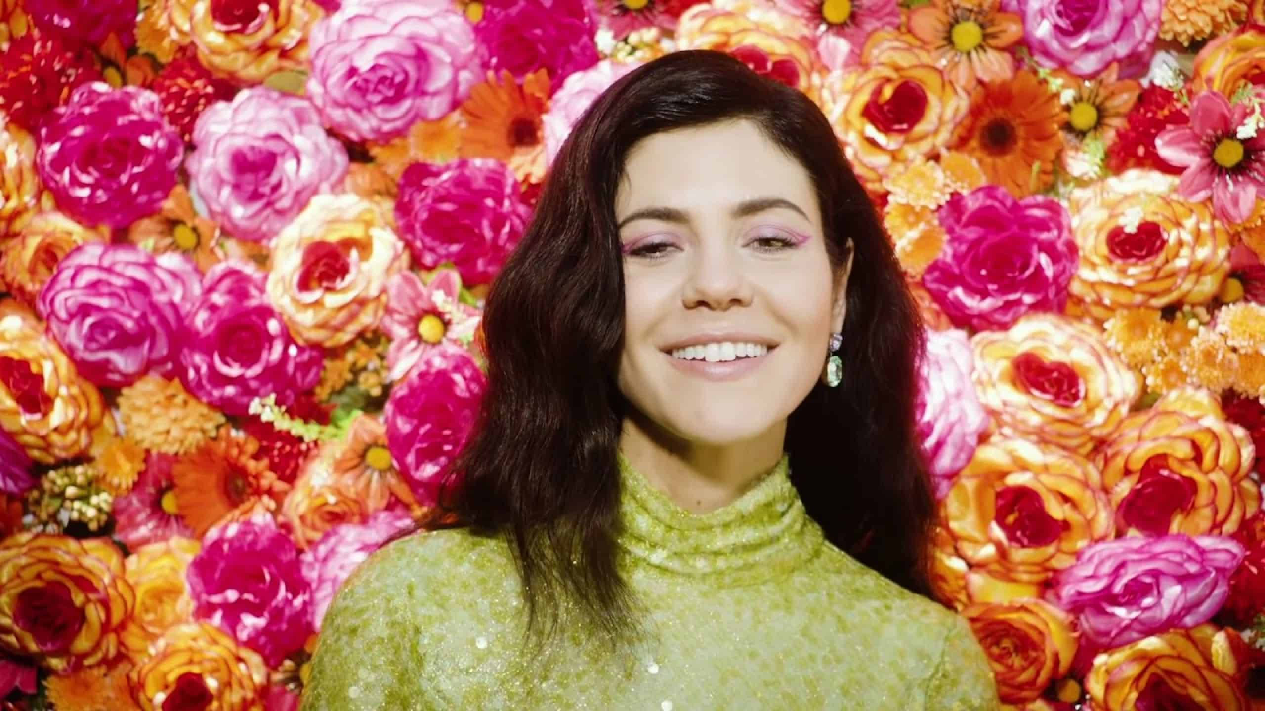 marina 桌布 壁紙 — 一位微笑的女性穿著閃亮綠色高領毛衣，站在充滿活力的粉紅和橙色玫瑰牆前。