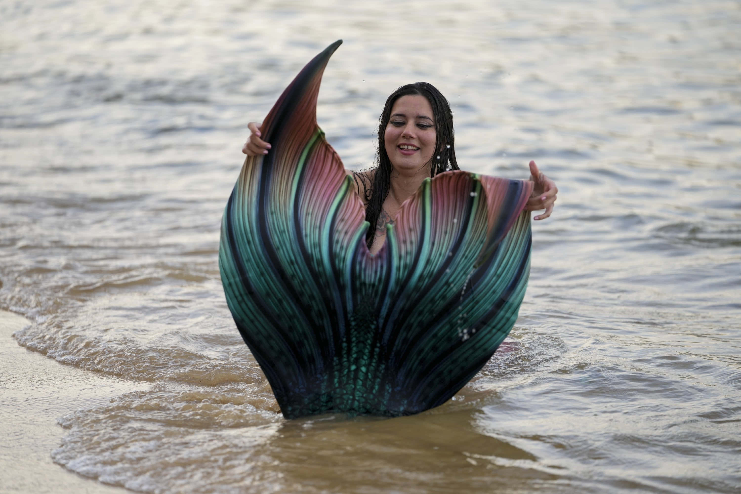 Mermaids 桌布 壁紙 — 一位微笑的女性穿著鮮豔的彩虹美人魚尾巴，站在夕陽下齊腰深的海浪中。