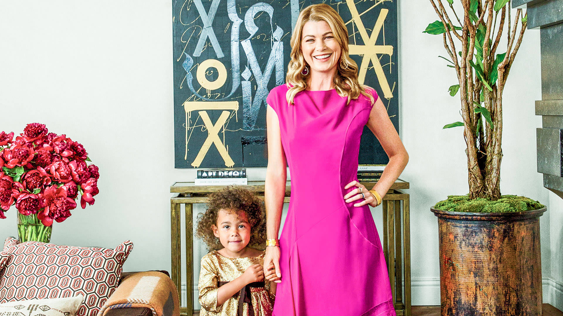 Ellen Pompeo 桌布 壁紙 — 一位微笑的女性穿著鮮豔的粉紅色連衣裙，與一個小孩站在時尚裝飾的客廳中。