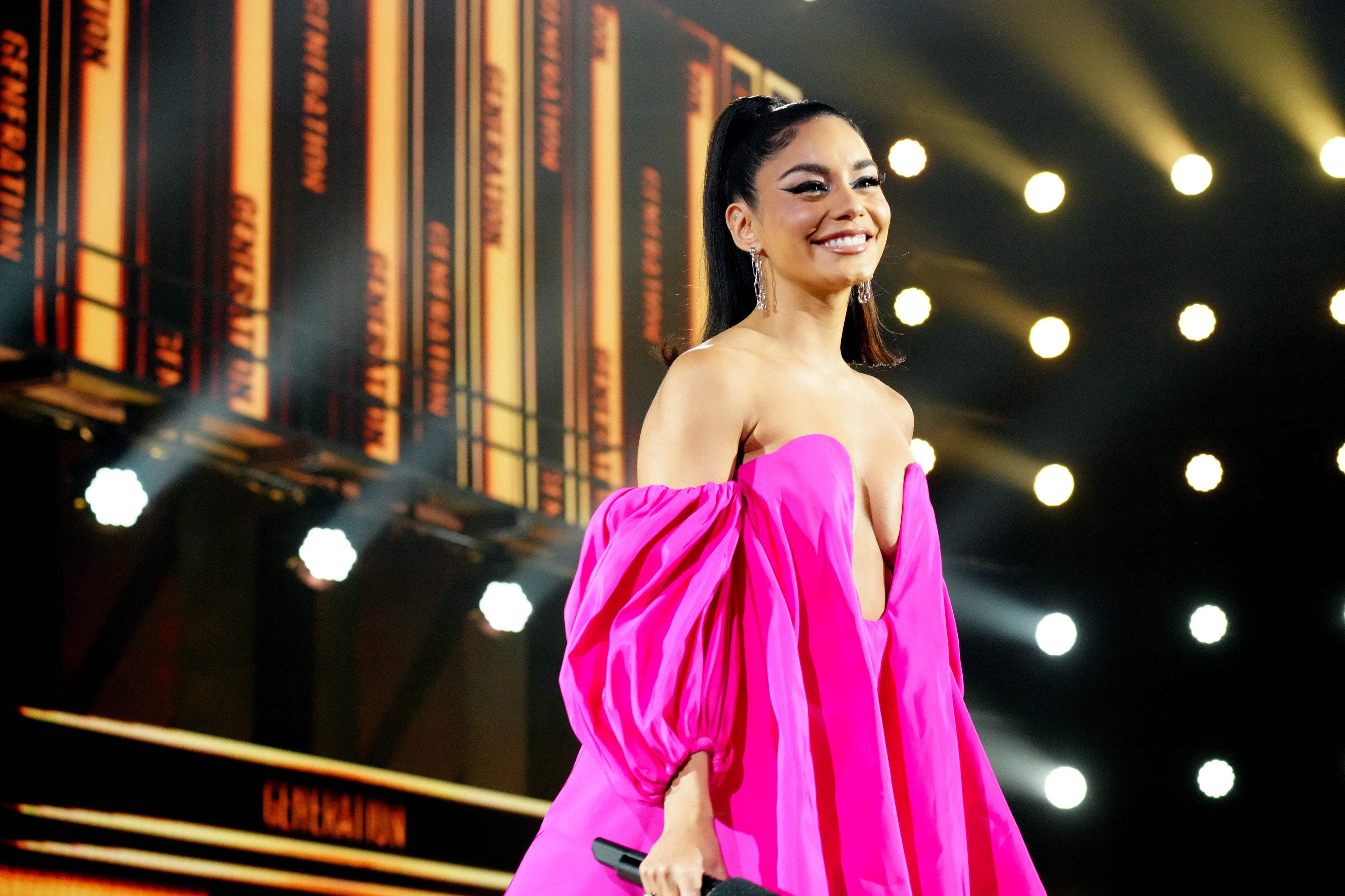 Celebrity 桌布 壁紙 — 一位微笑的女性穿著鮮豔的粉紅色露肩禮服，站在燈光璀璨的舞台上，身後有'GENERATION'標誌和聚光燈。