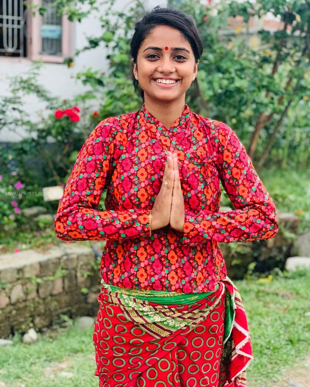 Nisha 桌布 壁紙 — 一位微笑的女性穿著鮮豔的紅色圖案傳統服裝，在戶外做出恭敬的問候手勢。
