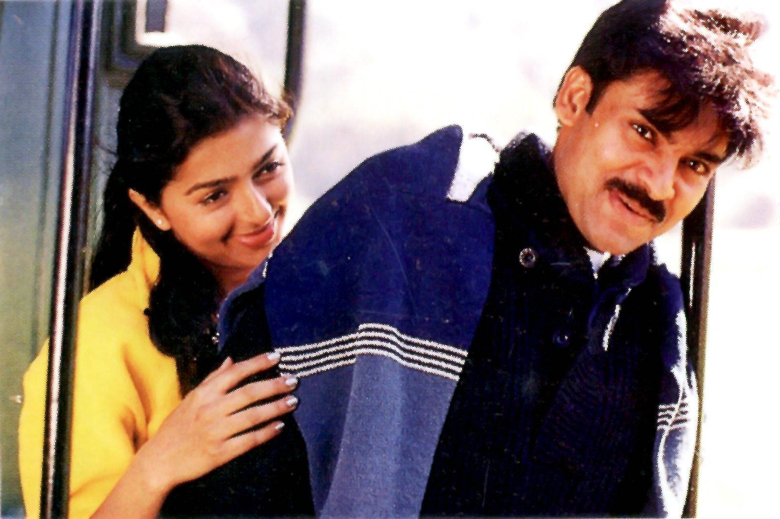 Pawan Kalyan 桌布 壁紙 — 一位微笑的女性穿著黃色上衣，深情地靠在一位穿著藍色夾克的男性身上，站在門口旁。