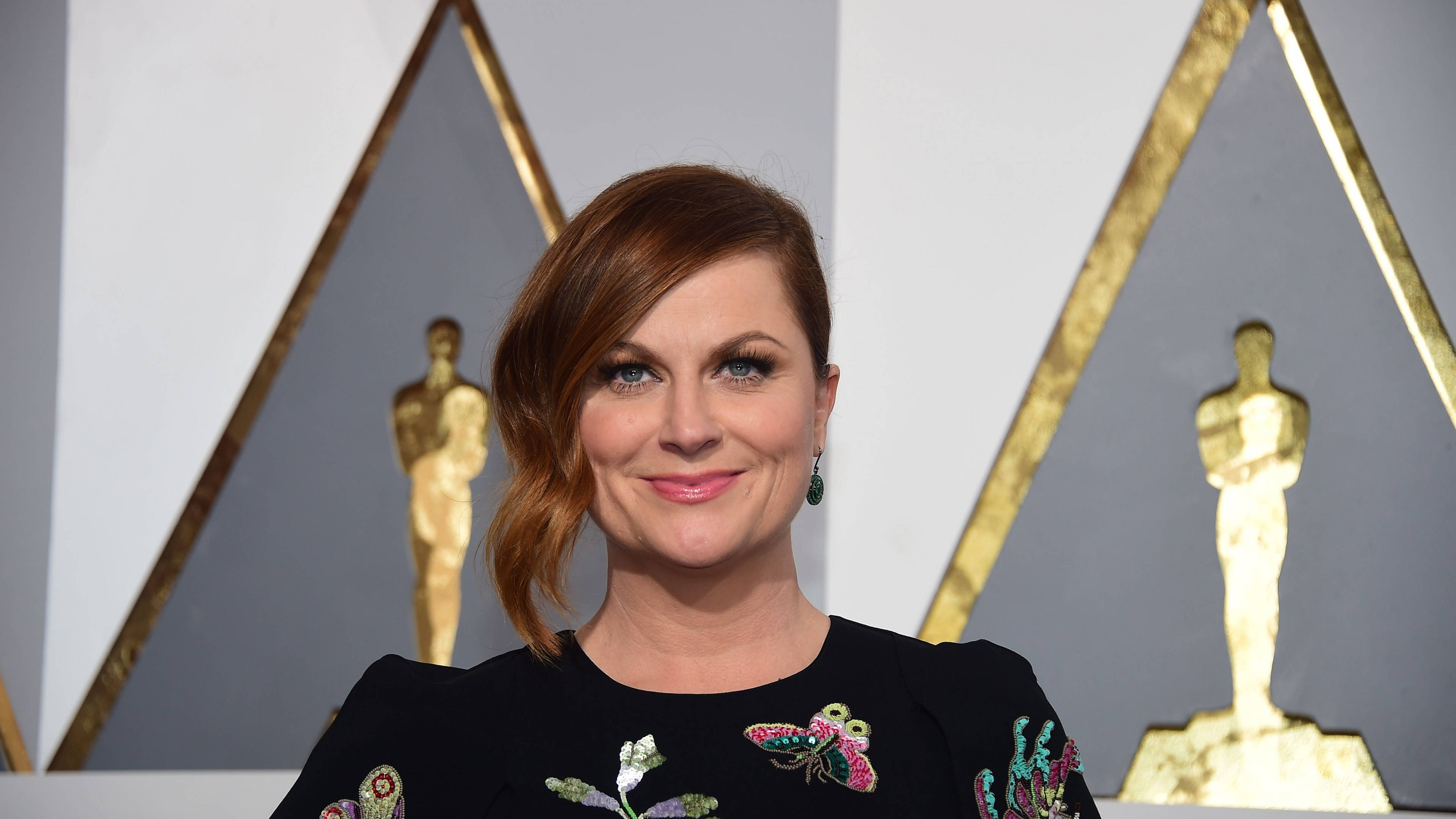 oscars 桌布 壁紙 — 一位微笑的女性穿著黑色刺繡連衣裙，在金色奧斯卡雕像和幾何金色邊框面板前擺姿勢。