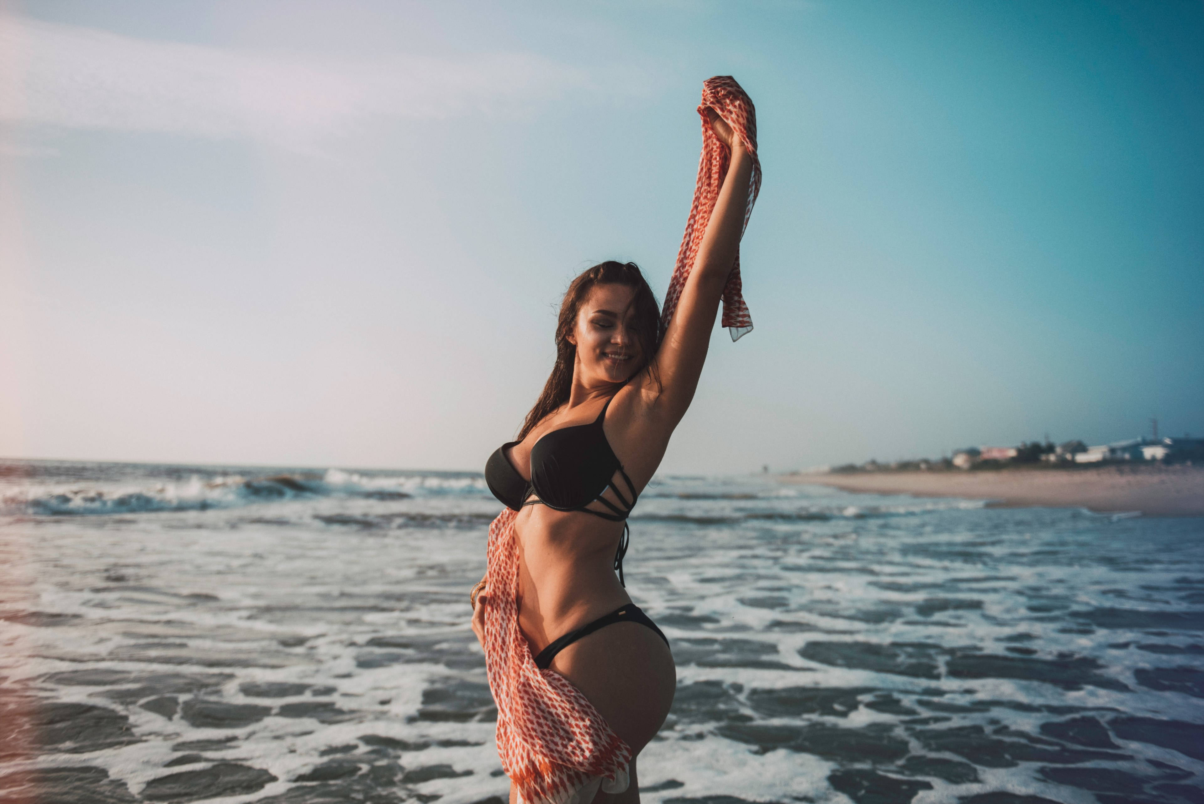海灘 桌布 壁紙 — 一位微笑的女性穿著黑色比基尼，在陽光明媚的海灘上舉起一條珊瑚圖案的毛巾，海浪拍打著沙灘。