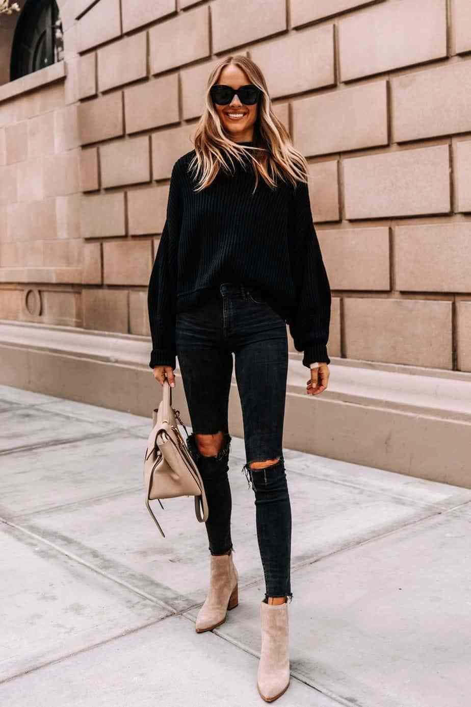 Black Clothing 桌布 壁紙 — 一位微笑的女性穿著黑色羅紋毛衣、破洞牛仔褲和米色靴子，在米色磚牆前的城市人行道上自信地行走。