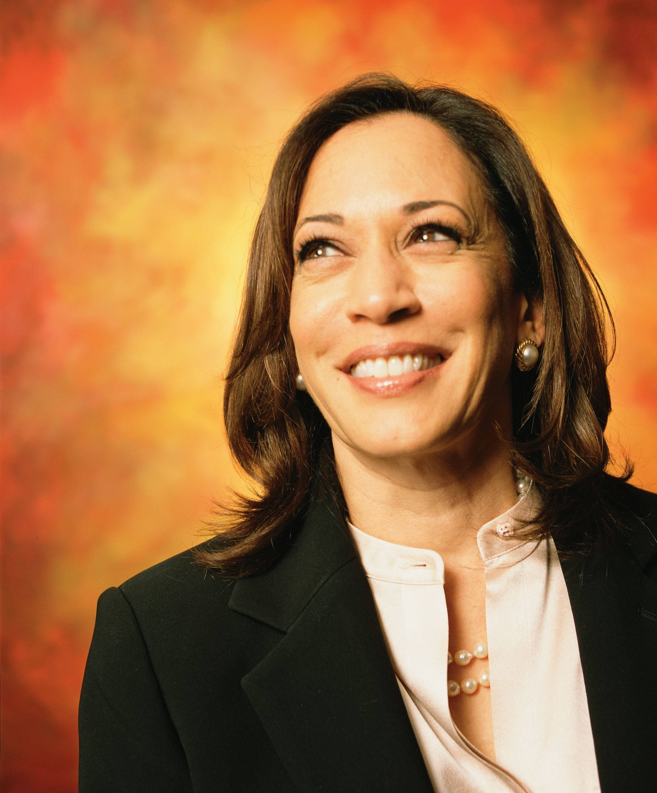 Kamala Harris 桌布 壁紙 — 一位微笑的女性穿著黑色西裝外套和珍珠項鍊，在溫暖的橙色和黃色漸變背景上向上看。