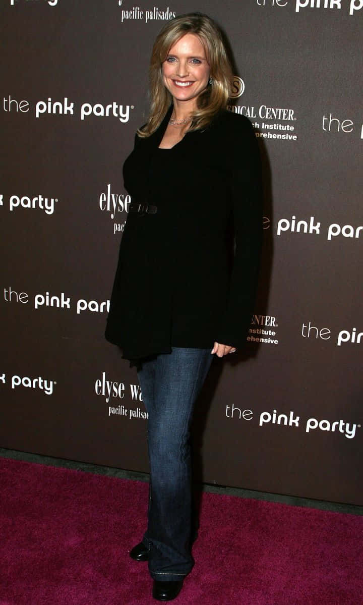 Courtney Thorne Smith 桌布 壁紙 — 一位微笑的女性穿著黑色西裝外套和牛仔褲，站在洋紅色地毯上，參加一個有品牌背景的粉紅色派對活動。