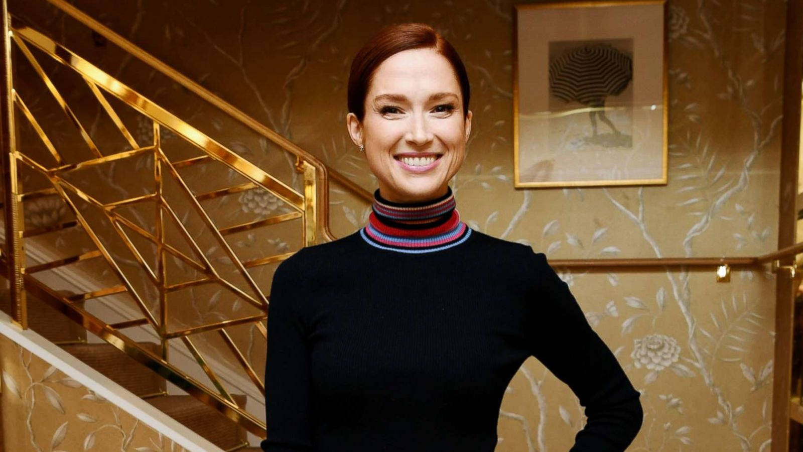 unbreakable kimmy schmidt 桌布 壁紙 — 一位微笑的女性穿著黑色高領衫搭配條紋領口，自信地站在華麗室內的金色樓梯旁。