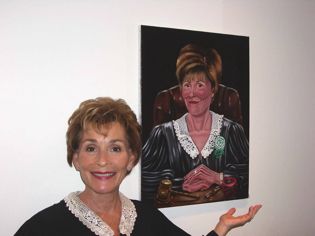 Judge Judy 桌布 壁紙 — 一位微笑的女性站在一幅自己作為法官的肖像畫旁邊，身穿帶有蕾絲領和綠色玫瑰胸針的黑色長袍。