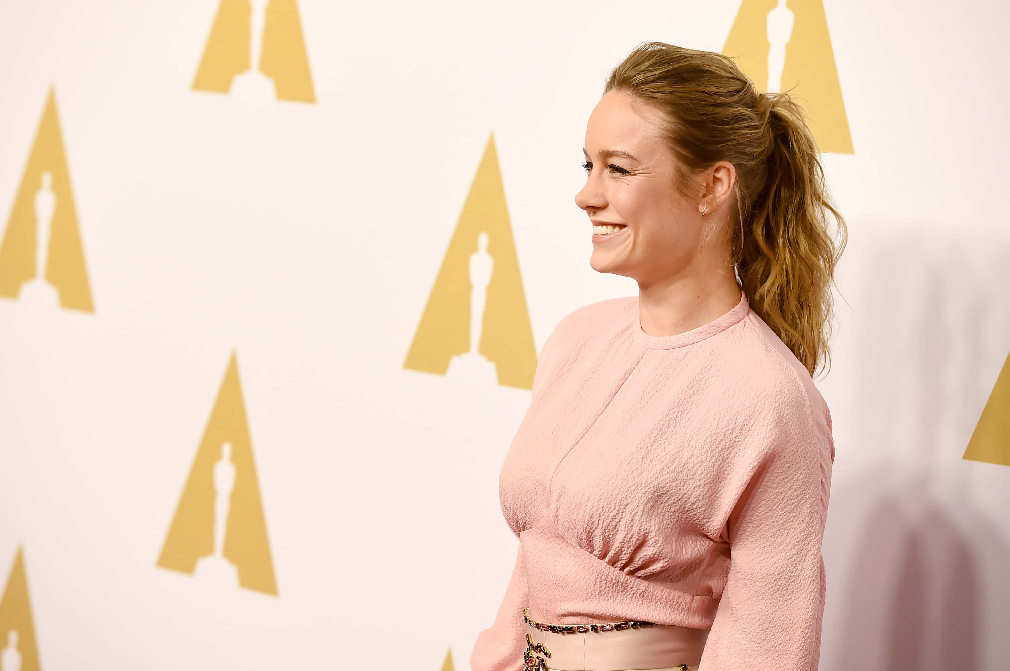 brie larson 桌布 壁紙 — 一位微笑的女性身穿粉紅色上衣，站在一面裝飾著重複金色奧斯卡小金人標誌的白色牆壁前。