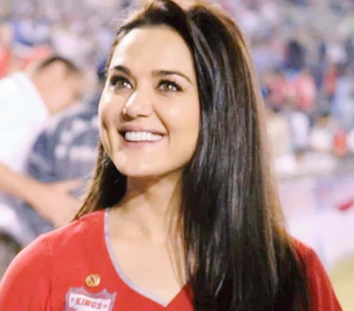 Preity Zinta 桌布 壁紙 — 一位微笑的女性身穿紅色 'Kings' 球衣，在身後體育場的人群中閃耀。