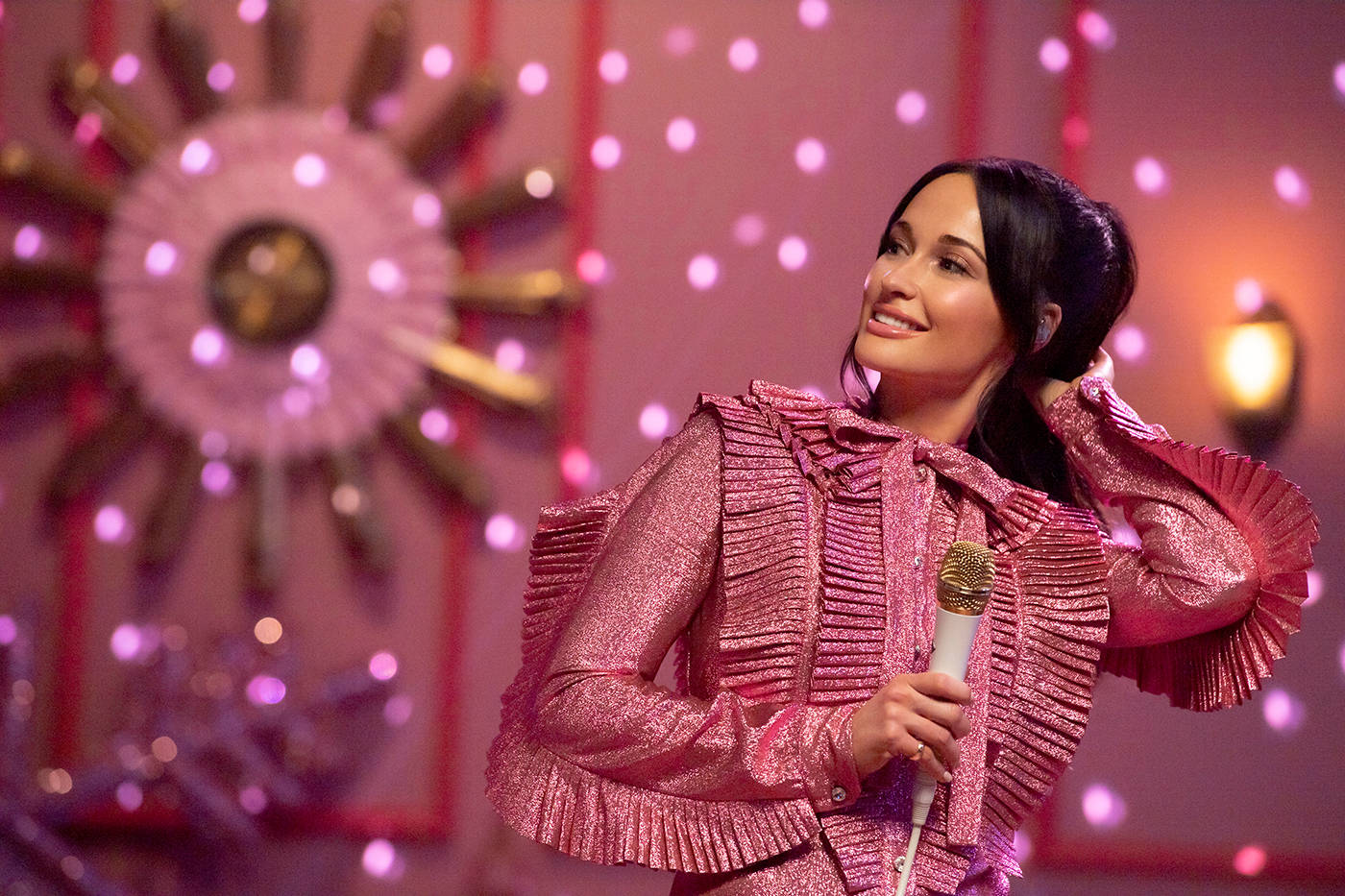 Kacey Musgraves 桌布 壁紙 — 一位微笑的女性身穿閃亮粉紅色荷葉邊外套，在裝飾著粉紅色燈光和大型裝飾太陽的舞台上握著麥克風。