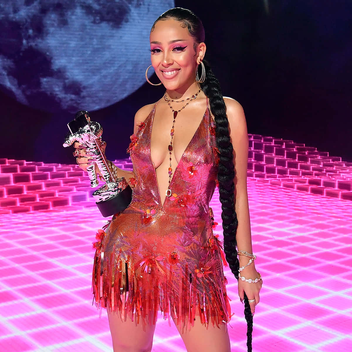 Doja Cat 桌布 壁紙 — 一位微笑的女性身穿閃亮紅色流蘇連衣裙，在發光的粉紅色網格舞台上手持獎盃，背景為月亮。