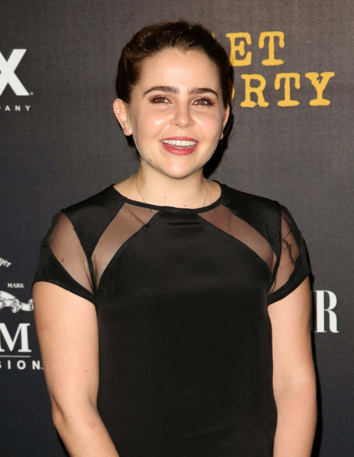 Mae Whitman 桌布 壁紙 — 一位微笑的女性身穿黑色透明袖連衣裙，在帶有品牌背景的紅地毯活動中擺姿勢。