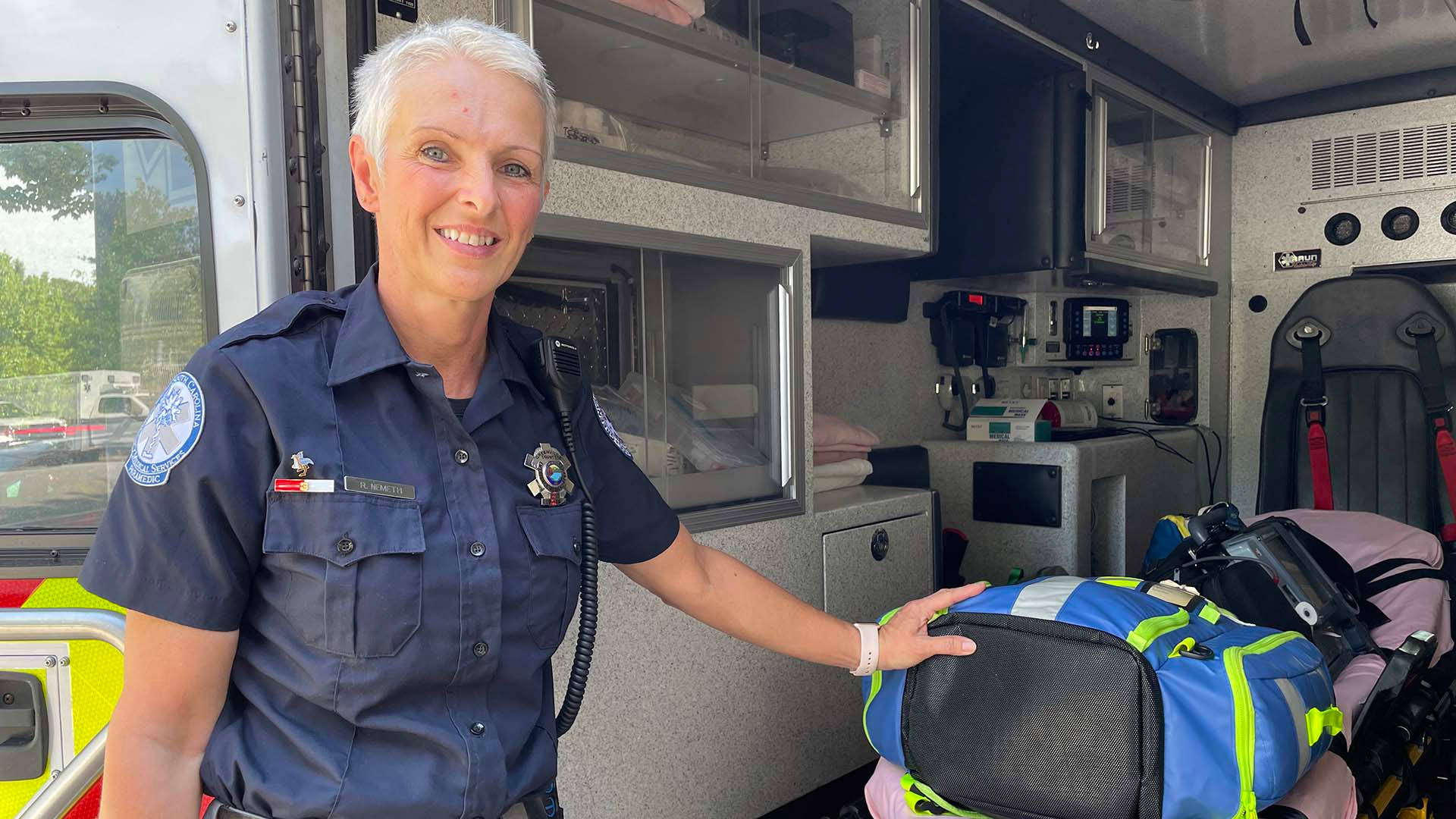 job 桌布 壁紙 — 一位微笑的女救護員穿著制服站在救護車內，手持醫療袋，周圍可見設備。