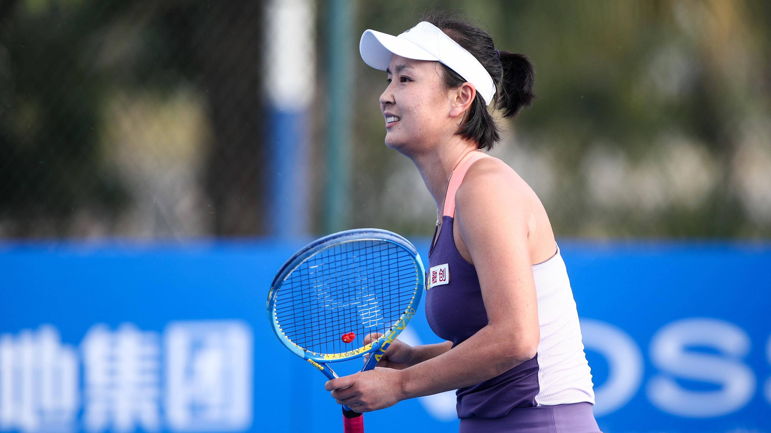Tennis 桌布 壁紙 — 一位微笑的女網球運動員，戴著白色遮陽帽，穿著紫色運動服，在藍色球場上拿著球拍，背景是模糊的綠色植物。