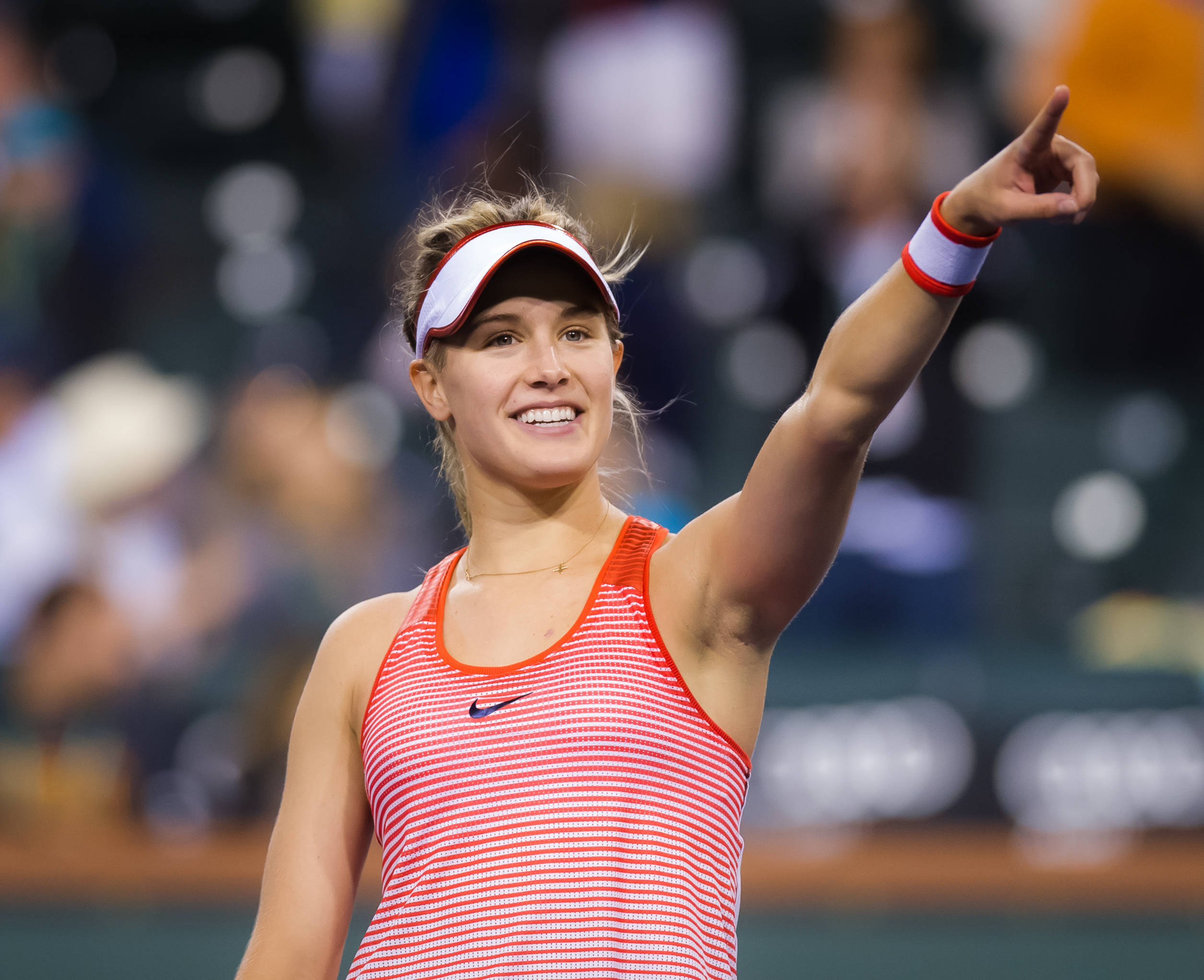 Eugenie Bouchard 桌布 壁紙 — 一位微笑的女網球運動員身穿紅色條紋背心和白色遮陽帽，在明亮的球場上高舉右臂指向天空慶祝勝利。