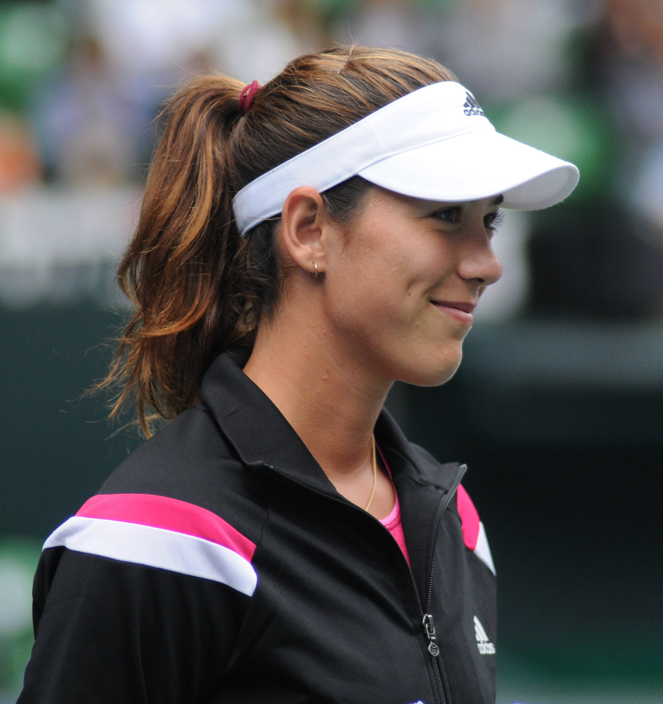 Tennis 桌布 壁紙 — 一位微笑的女網球選手，戴著白色遮陽帽，身穿帶有粉紅色點綴的黑色外套，在球場上看向旁邊。