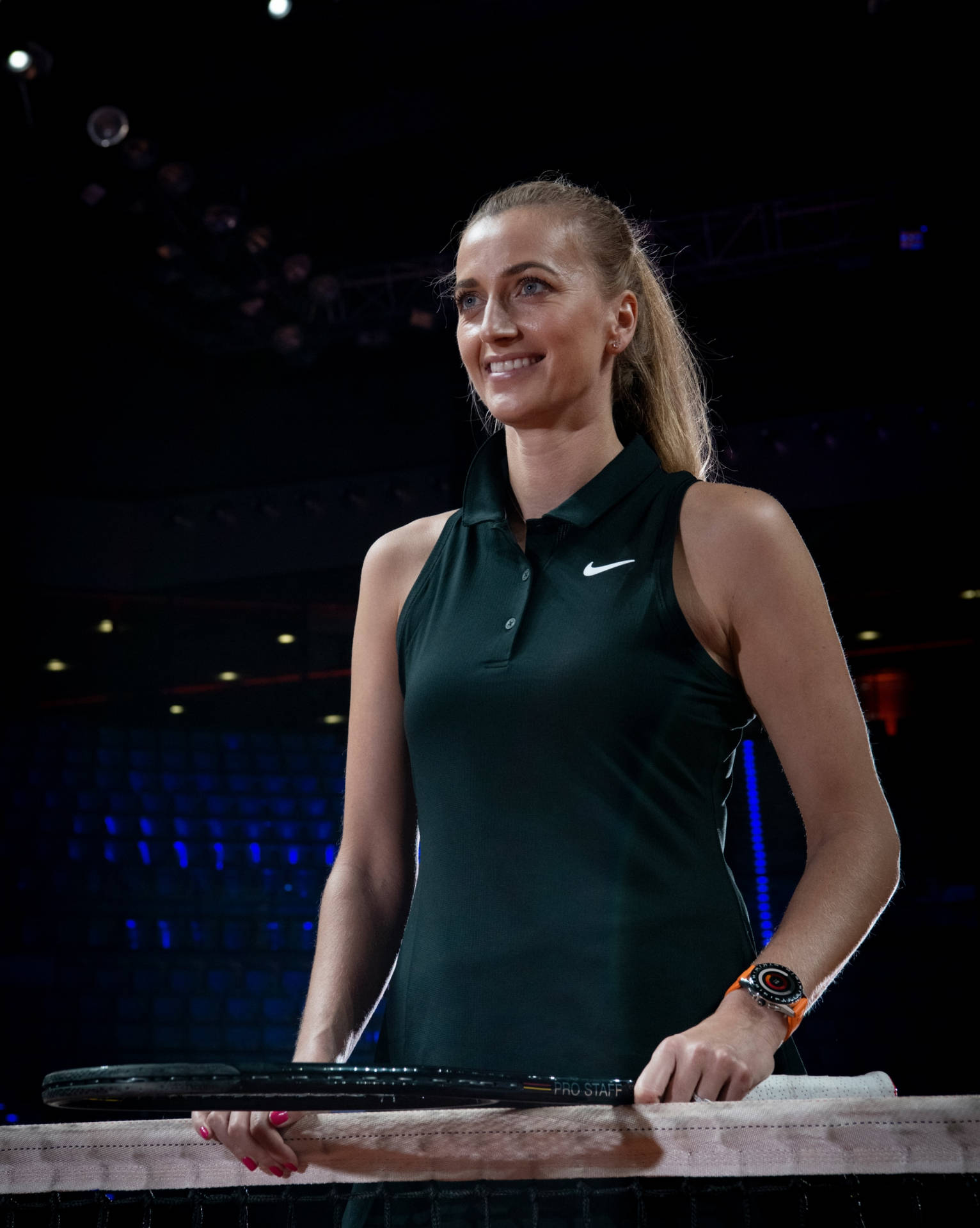 Tennis 桌布 壁紙 — 一位微笑的女網球選手穿著深綠色Polo衫，在明亮的體育場燈光下於球場上握著球拍。