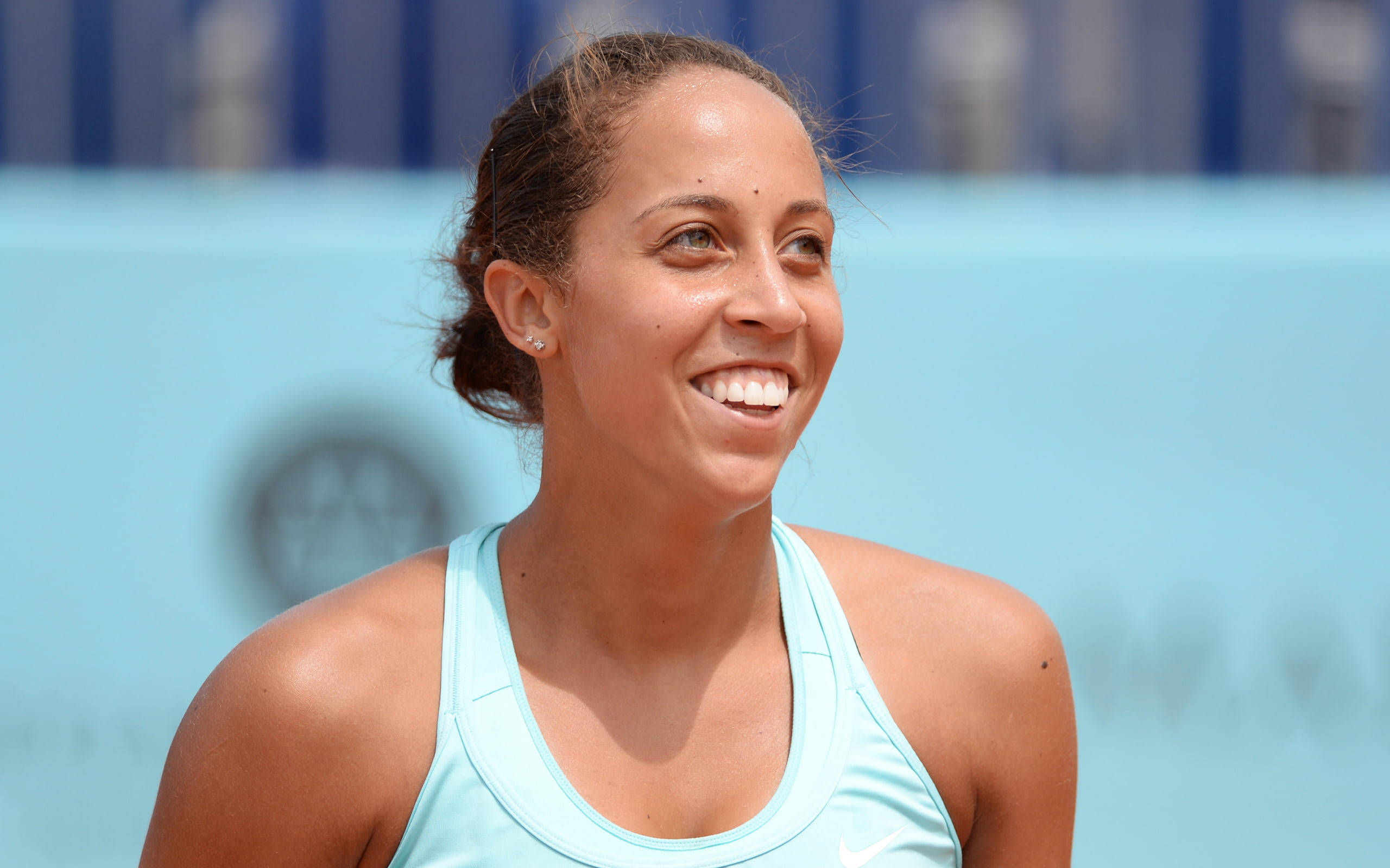 Athletics 桌布 壁紙 — 一位微笑的女網球選手穿著淺藍色背心，在明亮的陽光下看向鏡頭外，突顯她愉悅的表情。