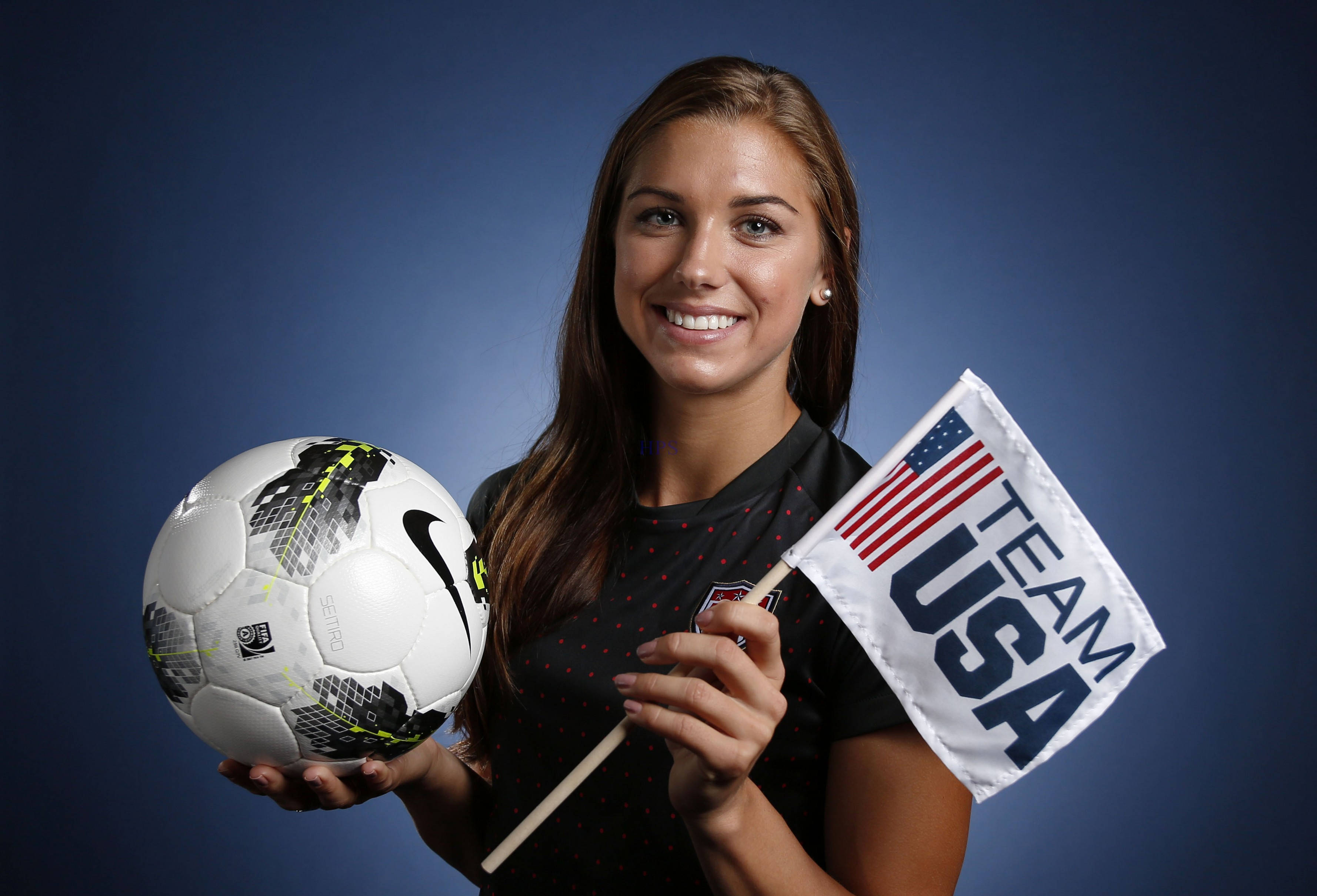 Alex Morgan 桌布 壁紙 — 一位微笑的女足球員在純藍色攝影棚背景前，手持Nike足球和Team USA旗幟。