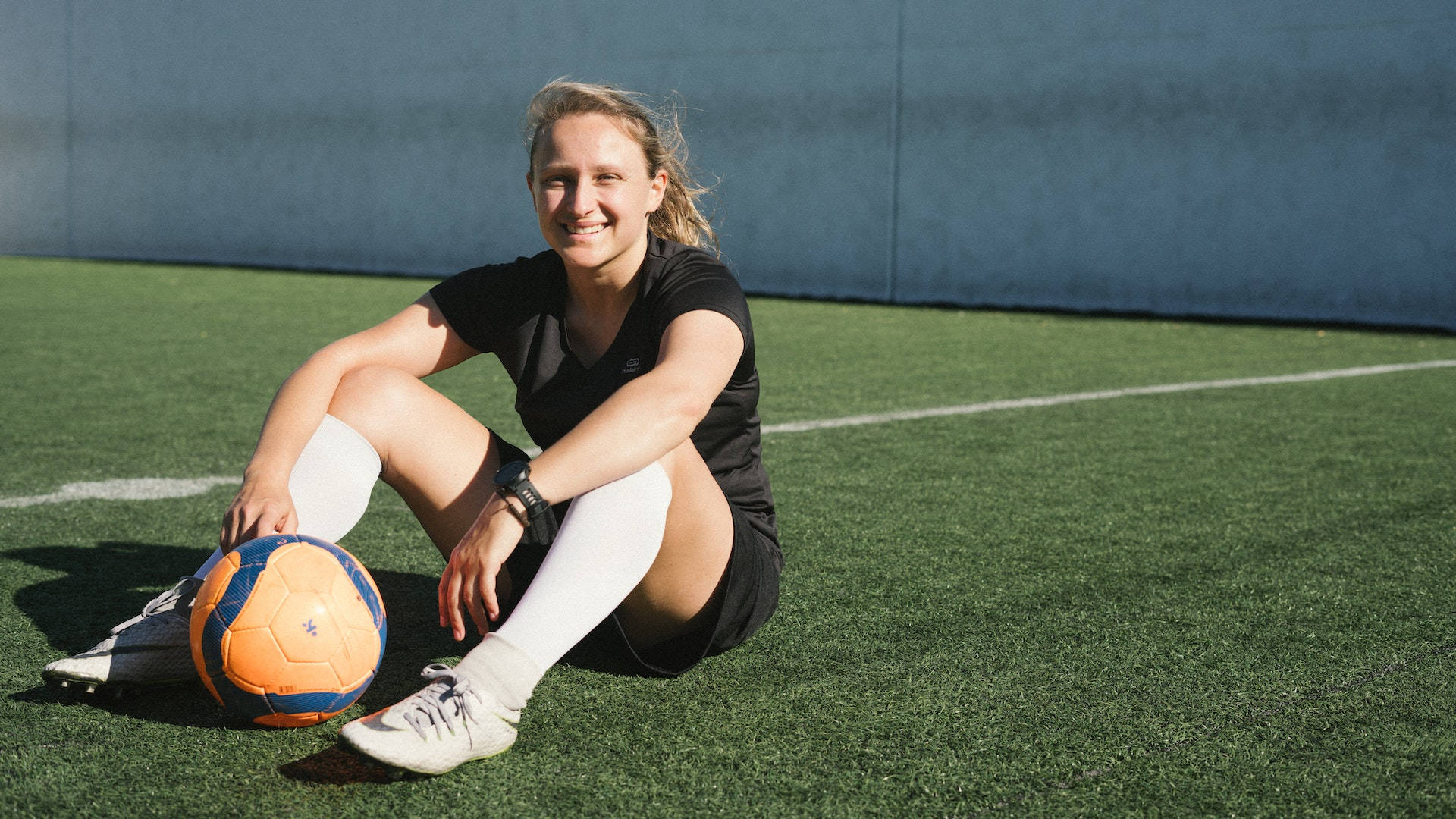 Athletic 桌布 壁紙 — 一位微笑的女足球員坐在綠色草地上，旁邊有一個橙色和藍色的足球，在明亮的陽光下。