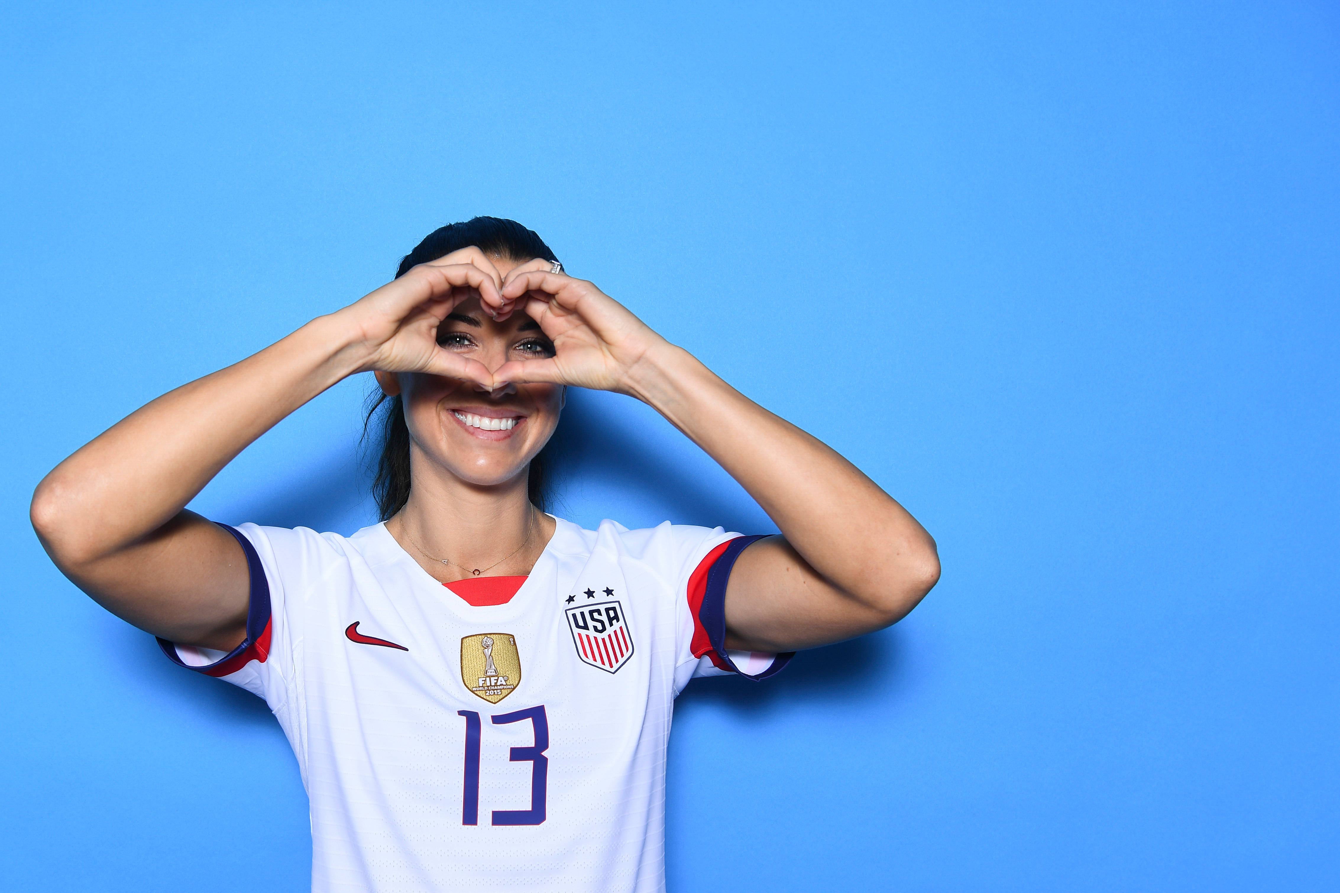 Alex Morgan 桌布 壁紙 — 一位微笑的女足球員身穿白色美國球衣，在純藍色背景前用手比出心形。