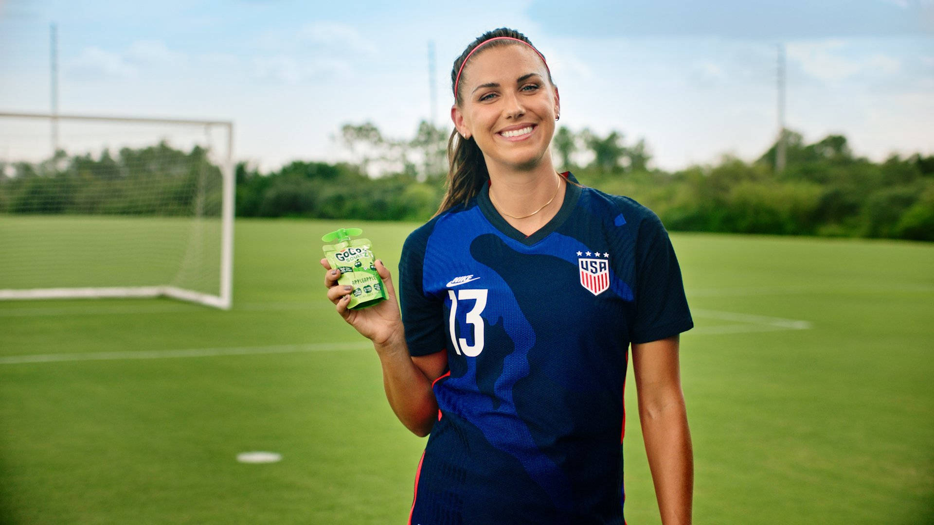 Alex Morgan 桌布 壁紙 — 一位微笑的女足球員身穿美國球衣，在陽光明媚的草地上，身後有球門網，手持綠色GoLo飲料。