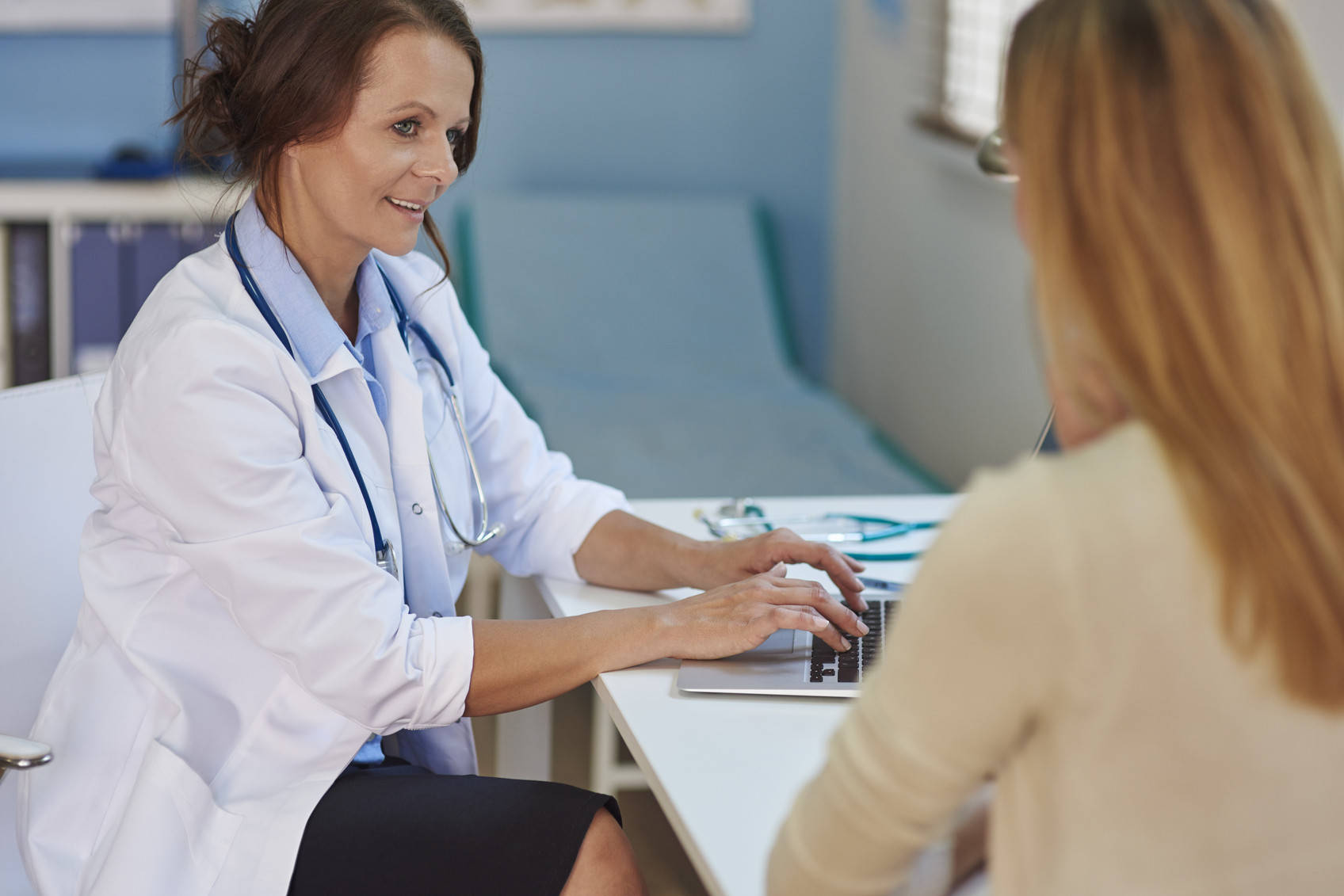 Hospital 桌布 壁紙 — 一位微笑的女醫生穿著白袍，在明亮的診間內一邊在筆記型電腦上打字，一邊與病人諮詢。