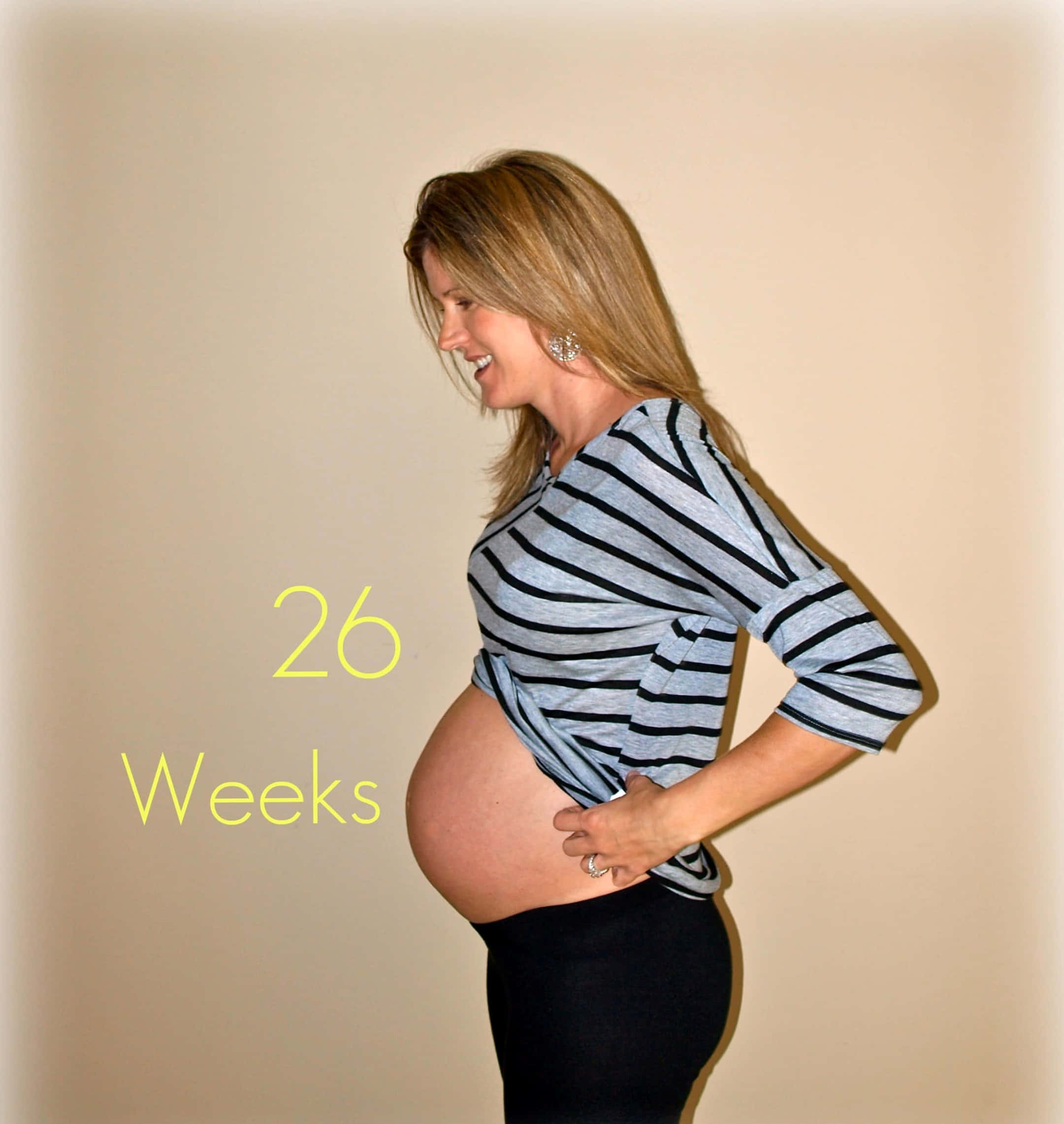 Pregnant 桌布 壁紙 — 一位微笑的孕婦穿著條紋襯衫，在柔和的米色工作室背景前自豪地展示她26週的肚子。