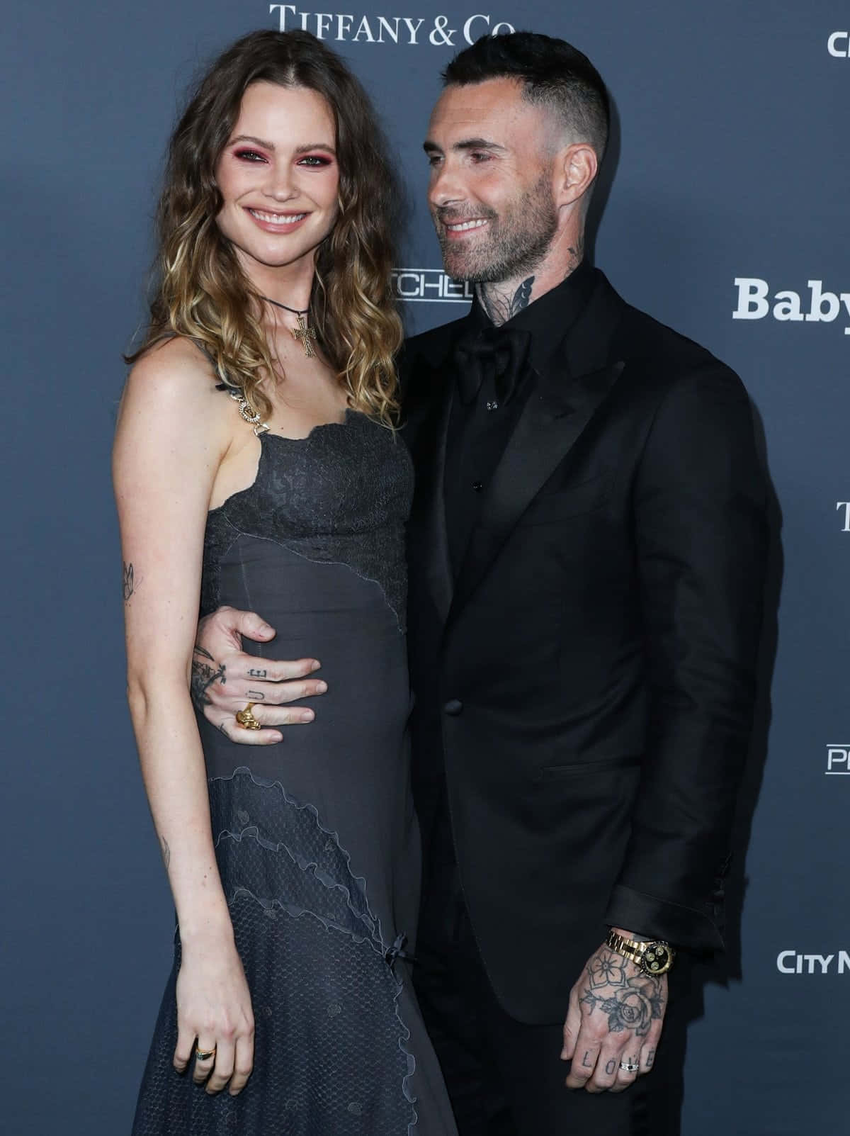 Beautiful Couple 桌布 壁紙 — 一位微笑的孕婦穿著深色蕾絲裙，站在一位紋身男子旁，他身穿黑色燕尾服，在一個品牌活動中。