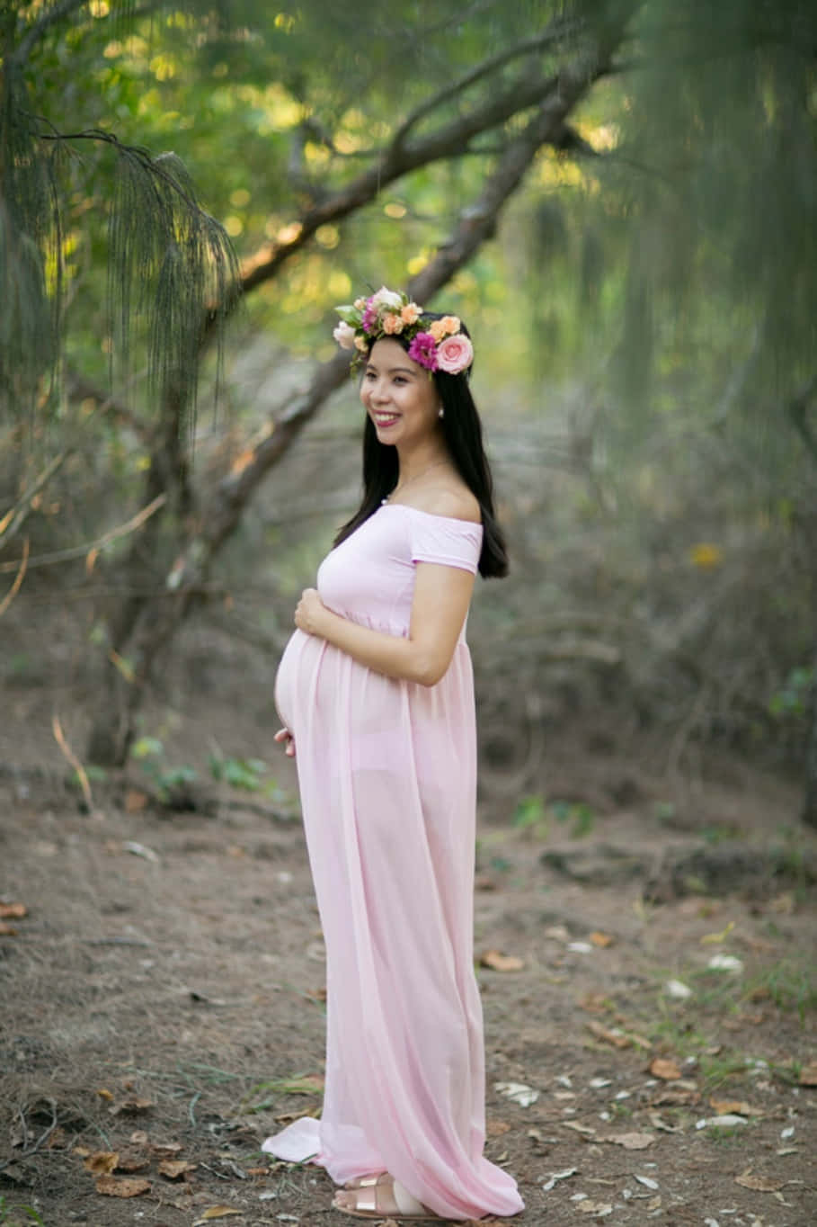 mother 桌布 壁紙 — 一位微笑的孕婦穿著飄逸的粉紅色連衣裙，站在陽光明媚的森林中，戴著色彩繽紛的花冠。
