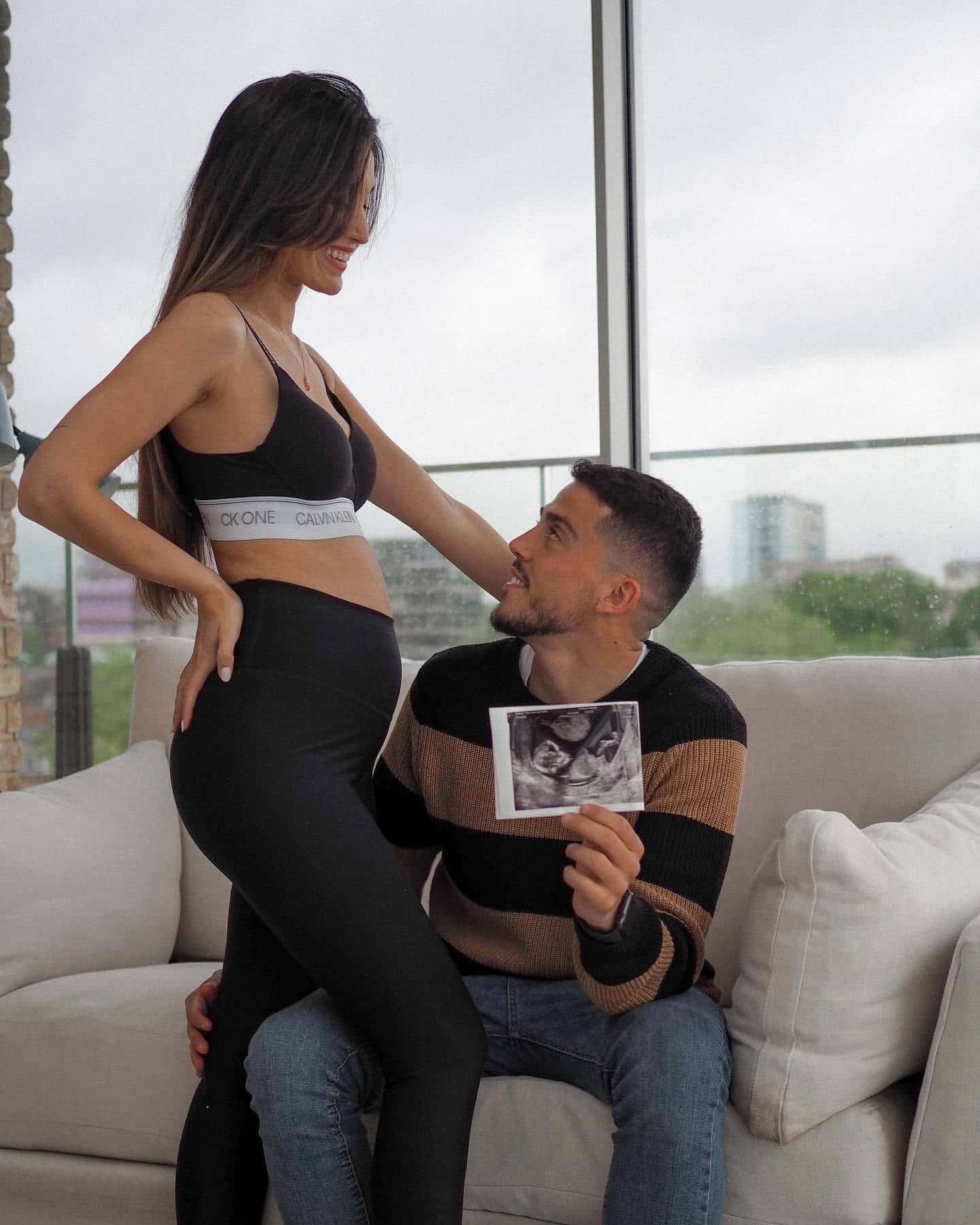 Pregnant 桌布 壁紙 — 一位微笑的孕婦站在她的伴侶上方，伴侶坐在沙發上拿著超音波照片，抬頭看著她。