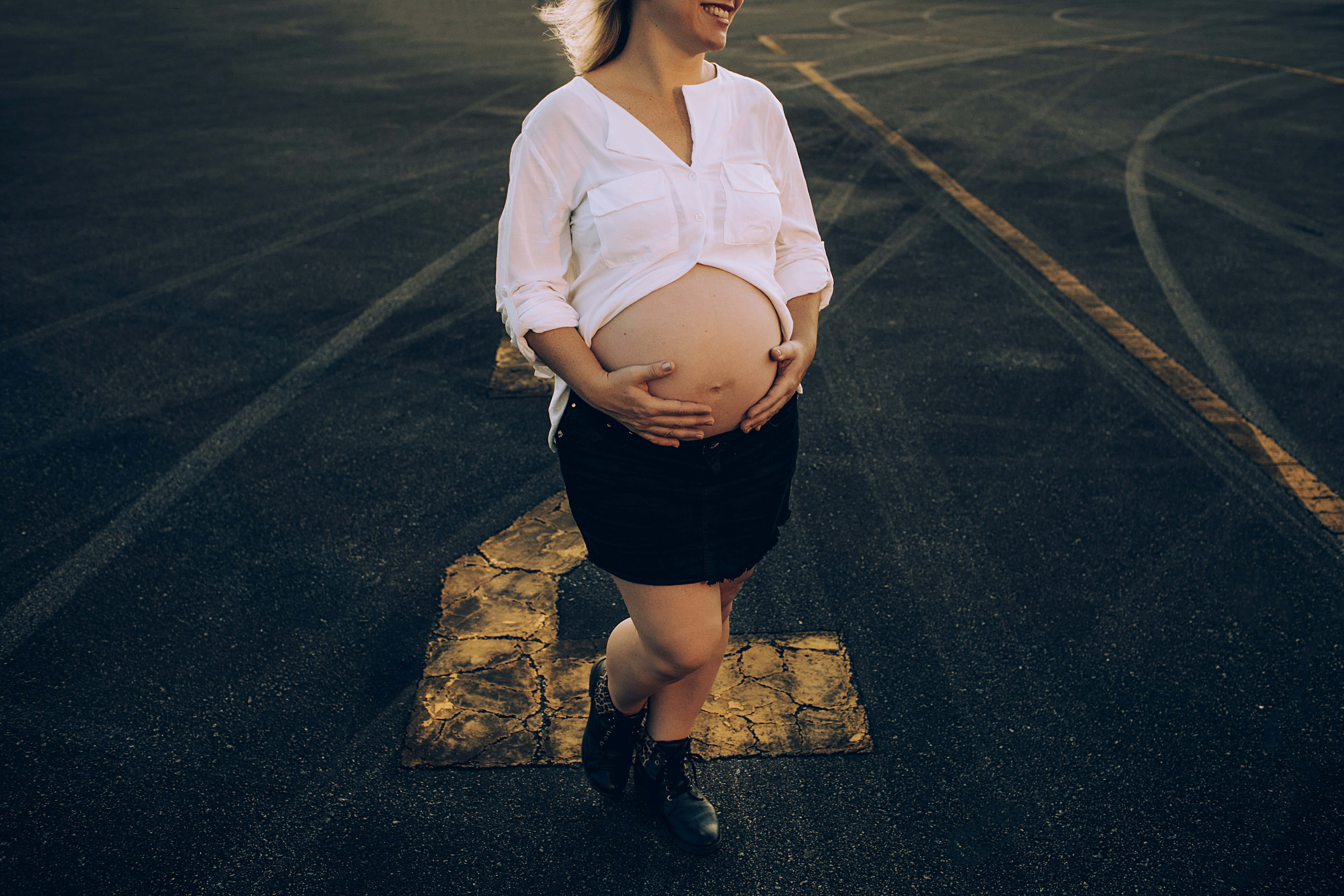 Pregnant 桌布 壁紙 — 一位微笑的孕婦身穿白襯衫和黑裙，站在裂開的瀝青停車場上，雙手輕捧腹部，沐浴在溫暖的金色光線中。