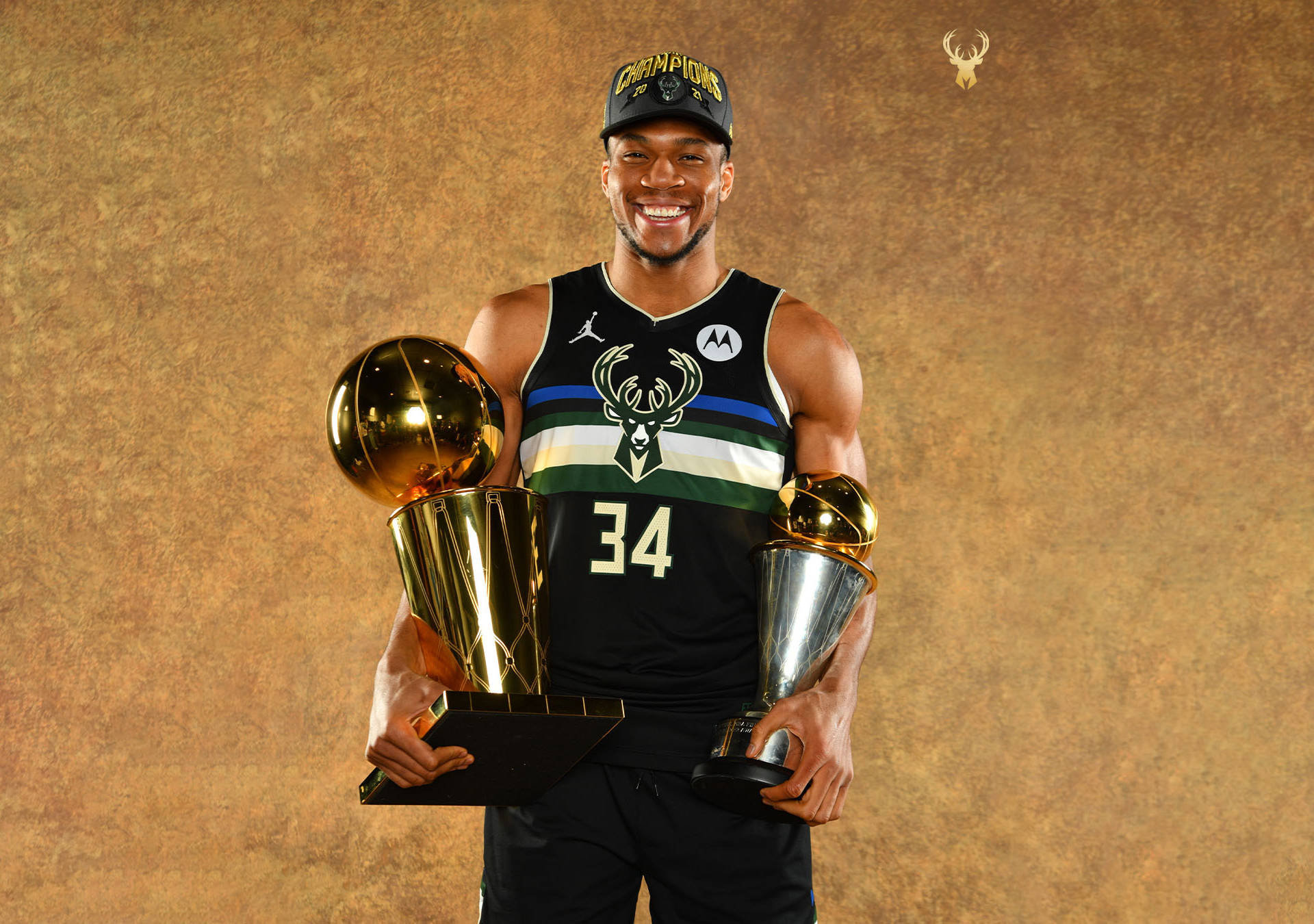 Champion 桌布 壁紙 — 一位微笑的密爾瓦基公鹿隊球員自豪地手持兩座閃閃發光的冠軍獎盃，背景是帶有紋理的金色背景。