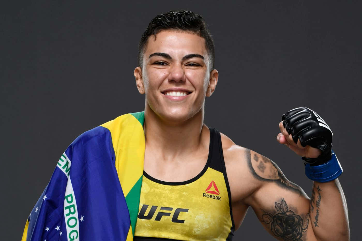 MMA 桌布 壁紙 — 一位微笑的巴西女性MMA拳手身穿UFC裝備，披著巴西國旗，自豪地舉起戴手套的拳頭。