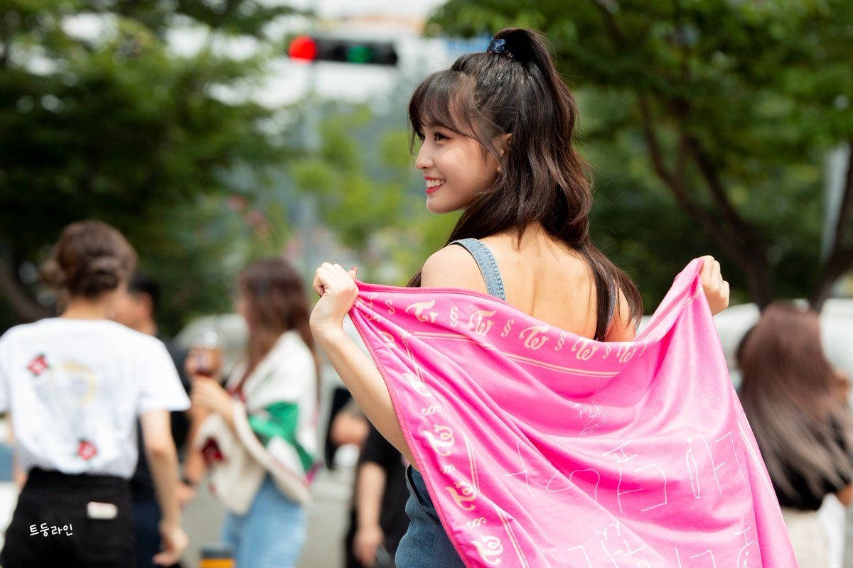 Momo 桌布 壁紙 — 一位微笑的年輕女子在城市街道上，回頭舉起一條帶有白色字樣的亮粉色圍巾。