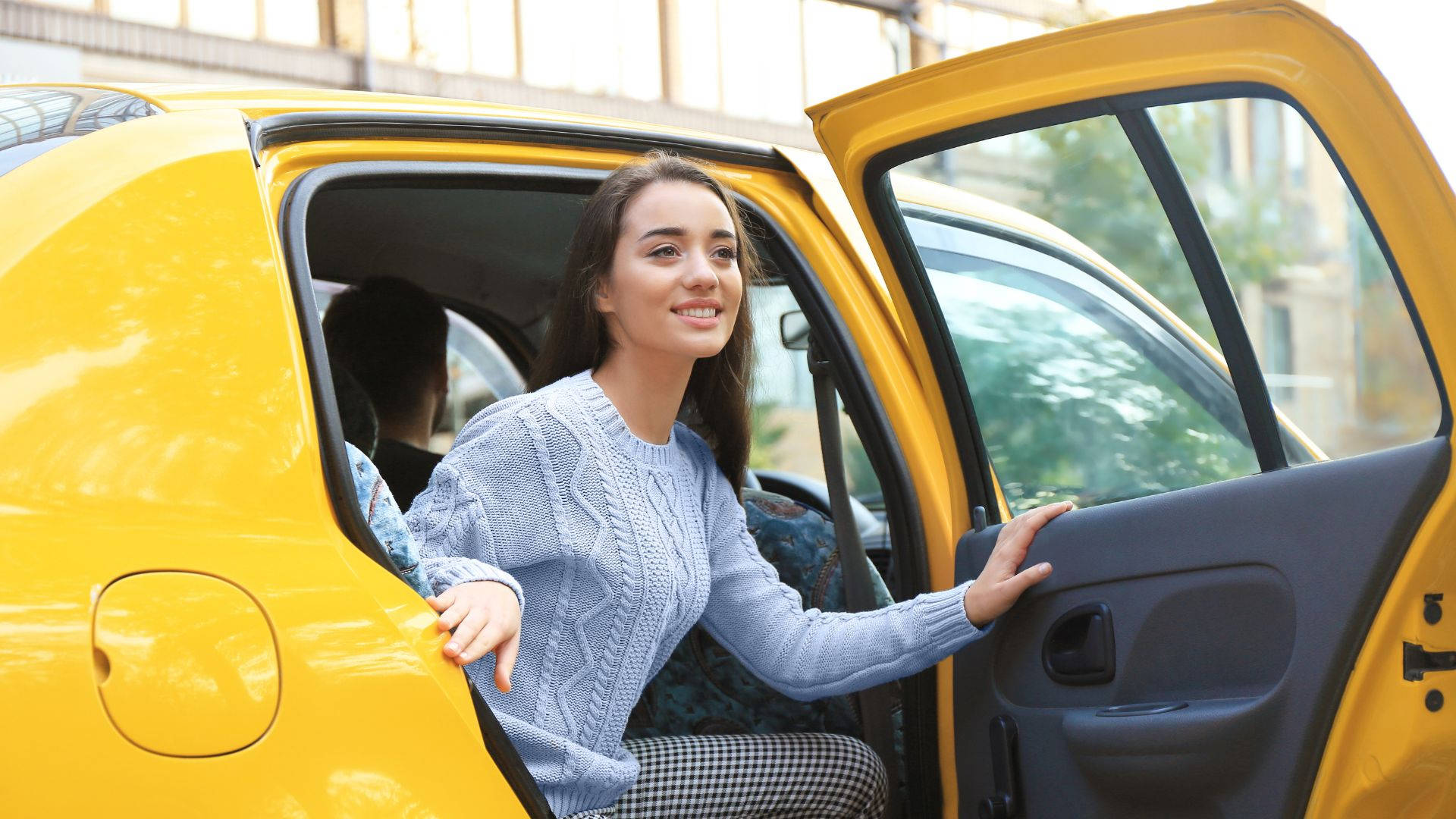 taxi 桌布 壁紙 — 一位微笑的年輕女子穿著淺藍色毛衣，在陽光明媚的日子裡從一輛亮黃色的車上下來。