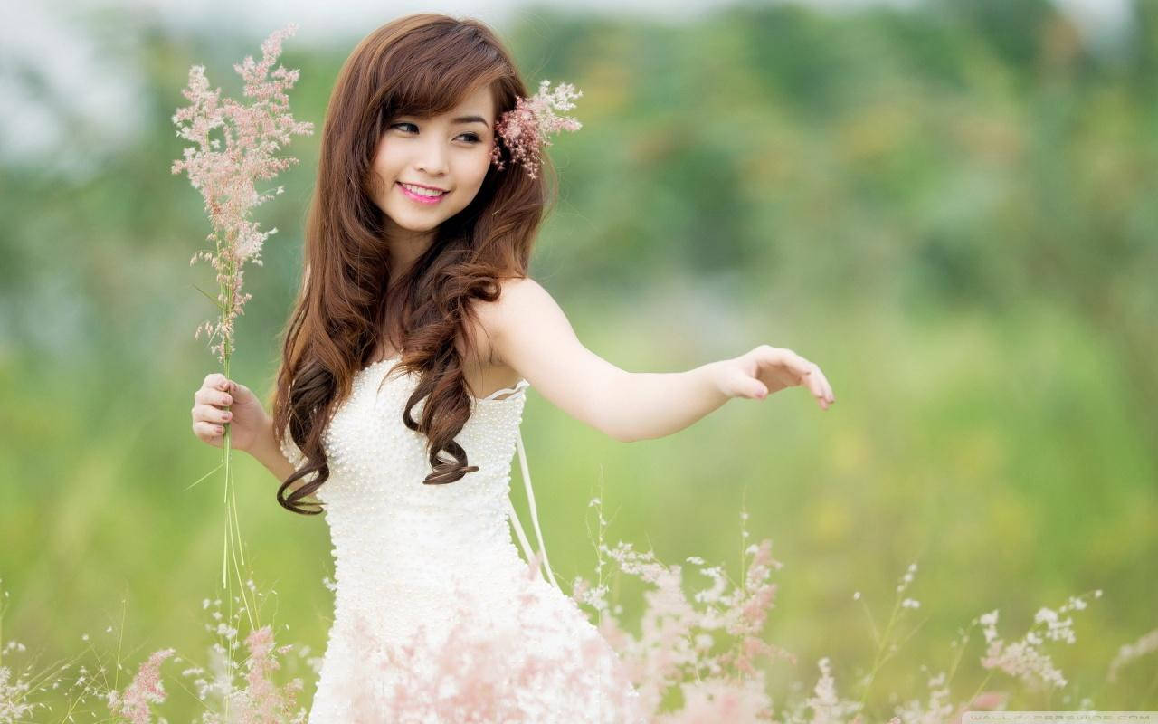 Woman 桌布 壁紙 — 一位微笑的年輕女子穿著白色連衣裙，在陽光明媚的綠色草地上手持粉紅色的草。