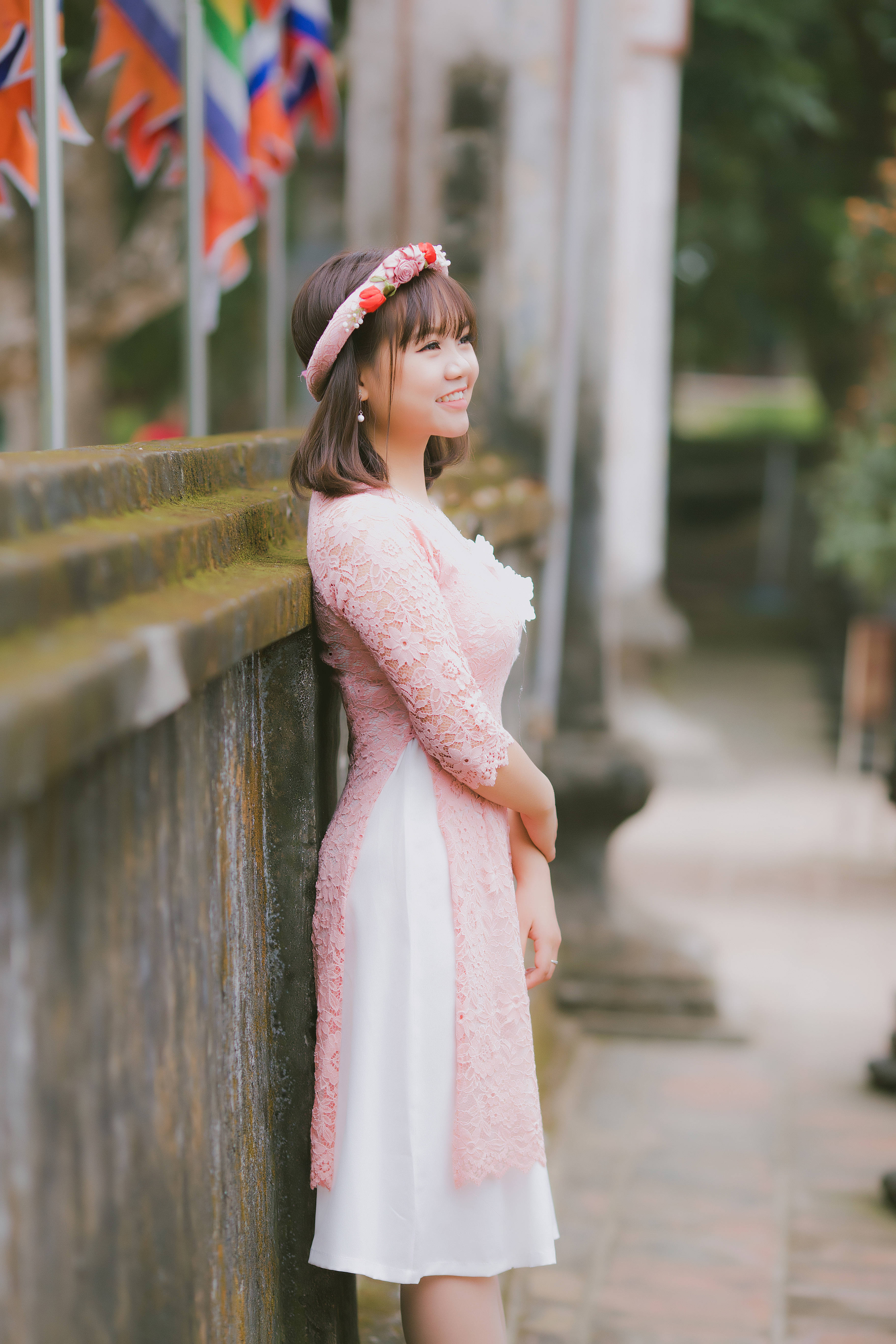 Cute Pink Girl 桌布 壁紙 — 一位微笑的年輕女子穿著粉紅色蕾絲連衣裙，在柔和日光下靠著石牆，背景有彩色旗幟。