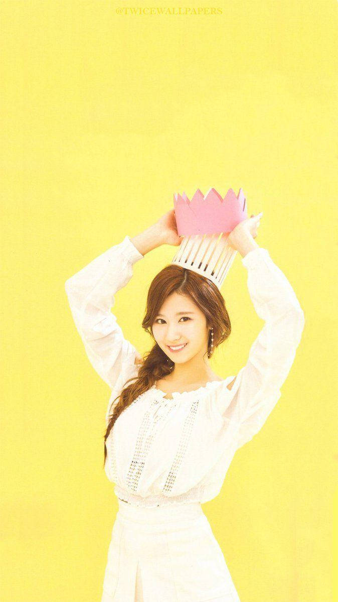 Twice Mina 桌布 壁紙 — 一位微笑的年輕女子身穿白色連衣裙，在明亮的黃色背景前，俏皮地將粉紅色紙皇冠舉過頭頂。