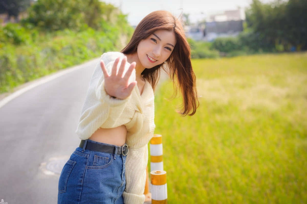 smiling 桌布 壁紙 — 一位微笑的年輕女子身穿米色毛衣和牛仔褲，在路邊護欄旁愉快地揮手，身後是茂密的綠草和蜿蜒的道路。