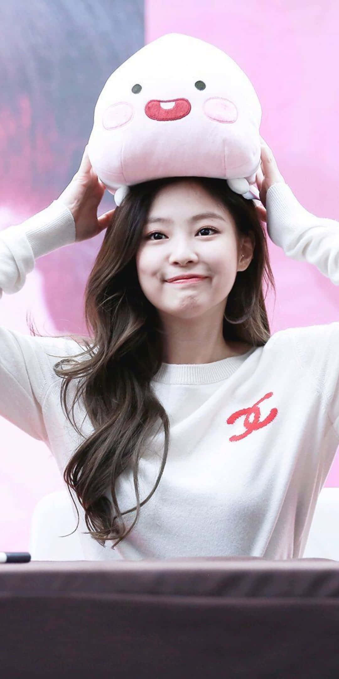 BLACKPINK 桌布 壁紙 — 一位微笑的年輕女性，穿著帶有紅色標誌的白色毛衣，頑皮地將一個粉紅色毛絨角色平衡在頭上。
