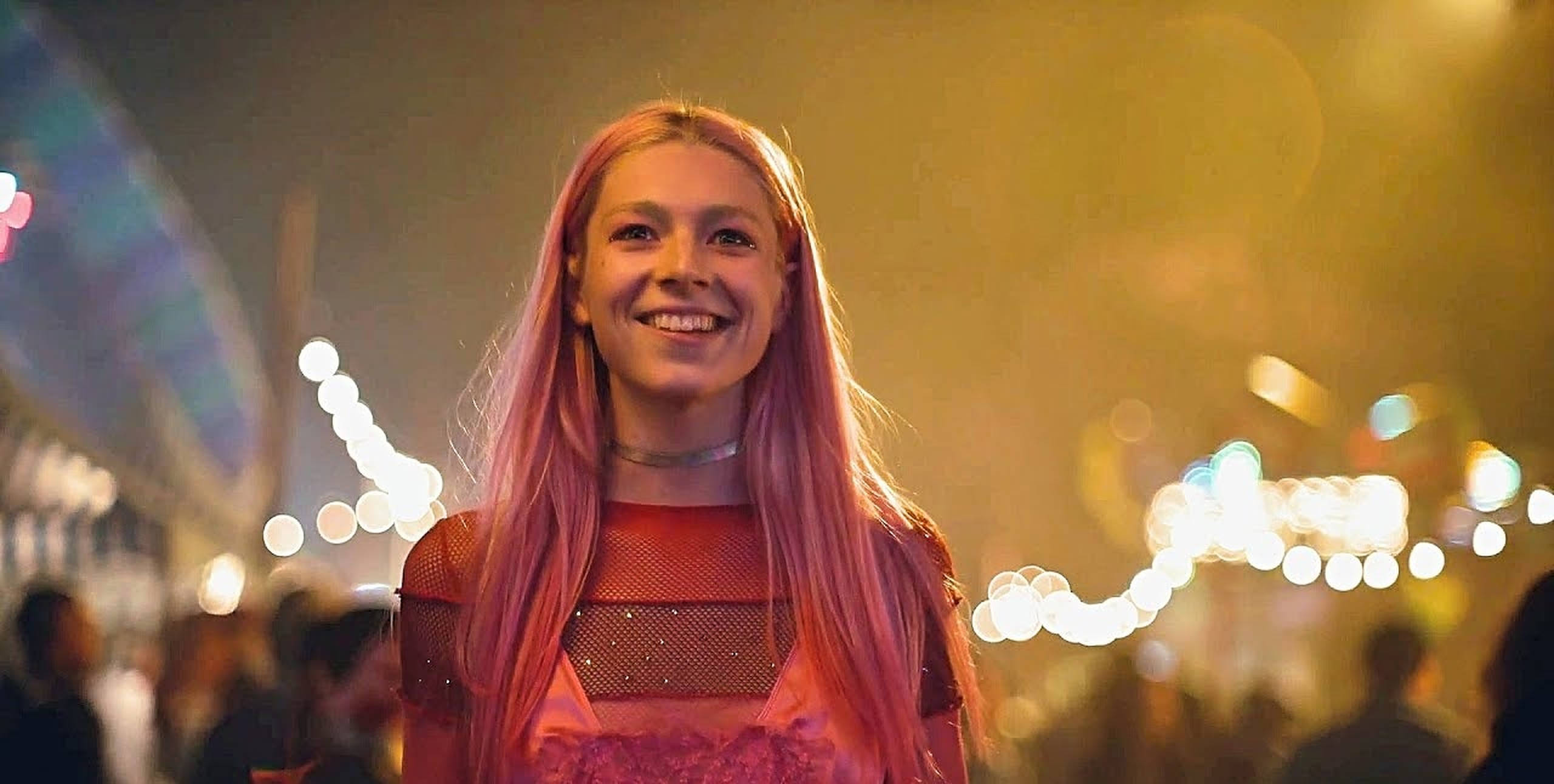 Hunter Schafer 桌布 壁紙 — 一位微笑的年輕女性，粉紅色頭髮，站在充滿活力的夜間嘉年華中，身後有發光的燈光和模糊的人群。